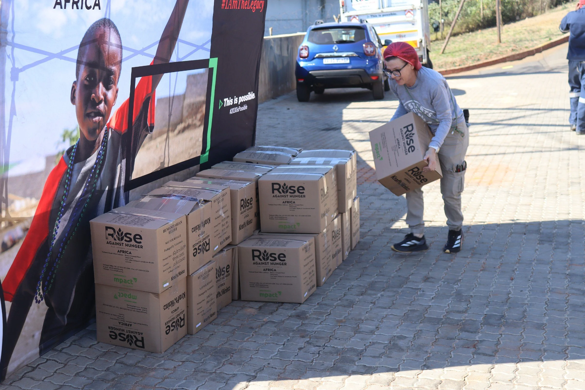 A woman wearing glasses, a red headband, and gray clothing is placing a cardboard box into a pile of similar boxes labeled "Rise Against Hunger Africa" on a cobblestone sidewalk. There is a large banner with a child's face and the hashtag #IAmTheLega