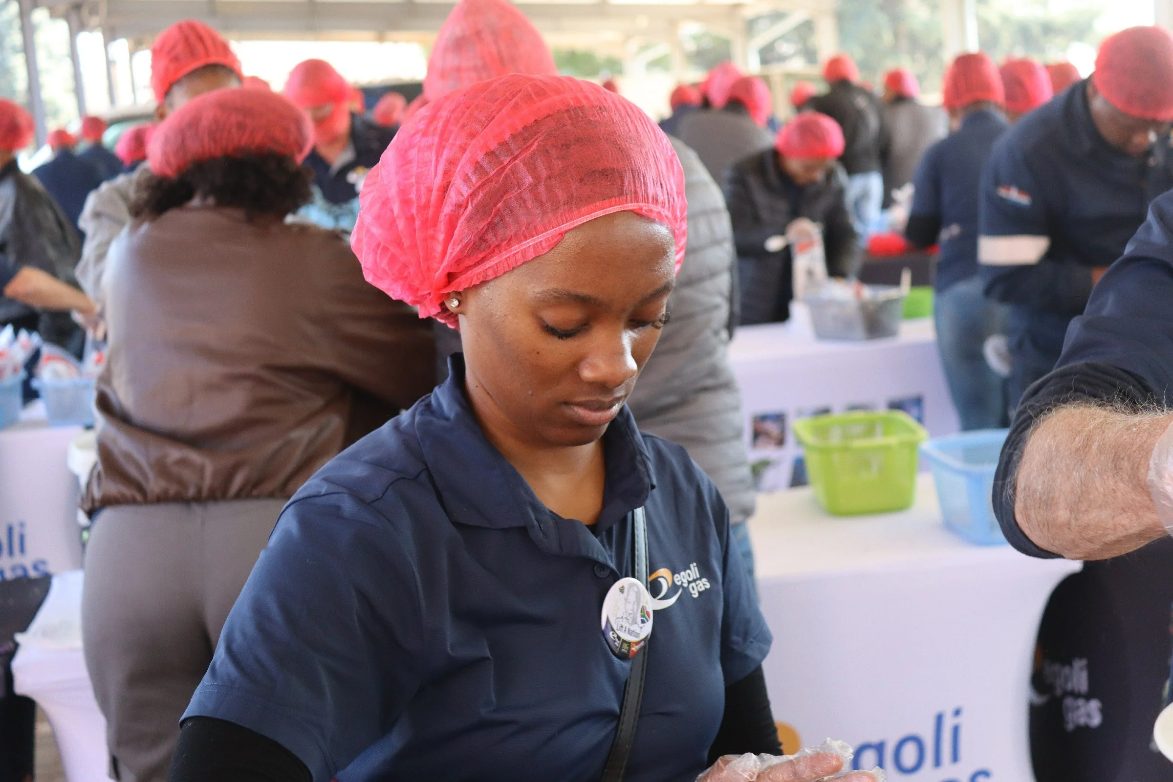 A group of people wearing pink hairnets participating in an event, with the focus on a woman in the foreground wearing a navy blue jacket and earrings, working at a table.