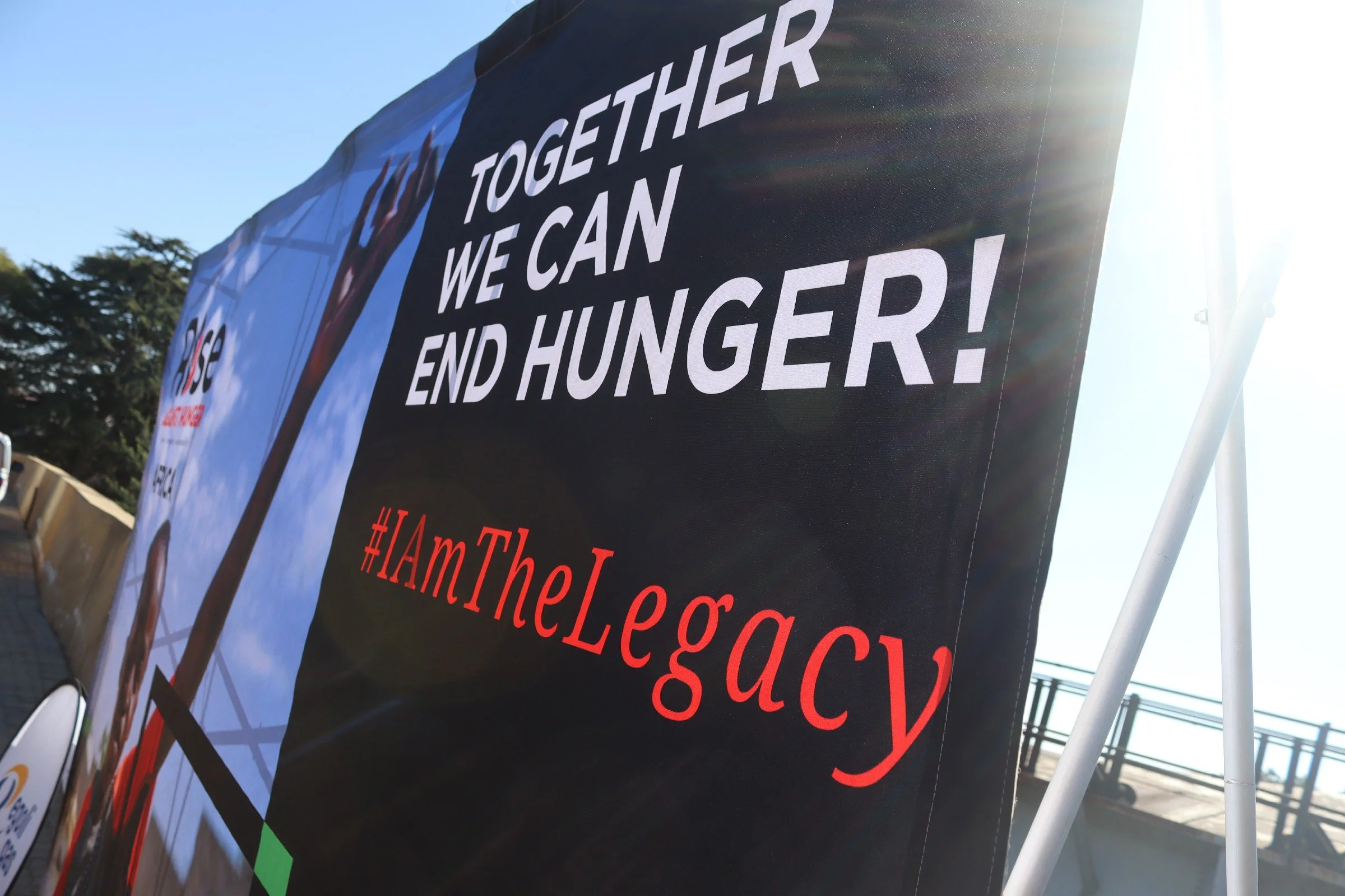 A black banner with white text reading 'Together We Can End Hunger!' is displayed outdoors, illuminated by sunlight, with trees and a clear blue sky in the background.