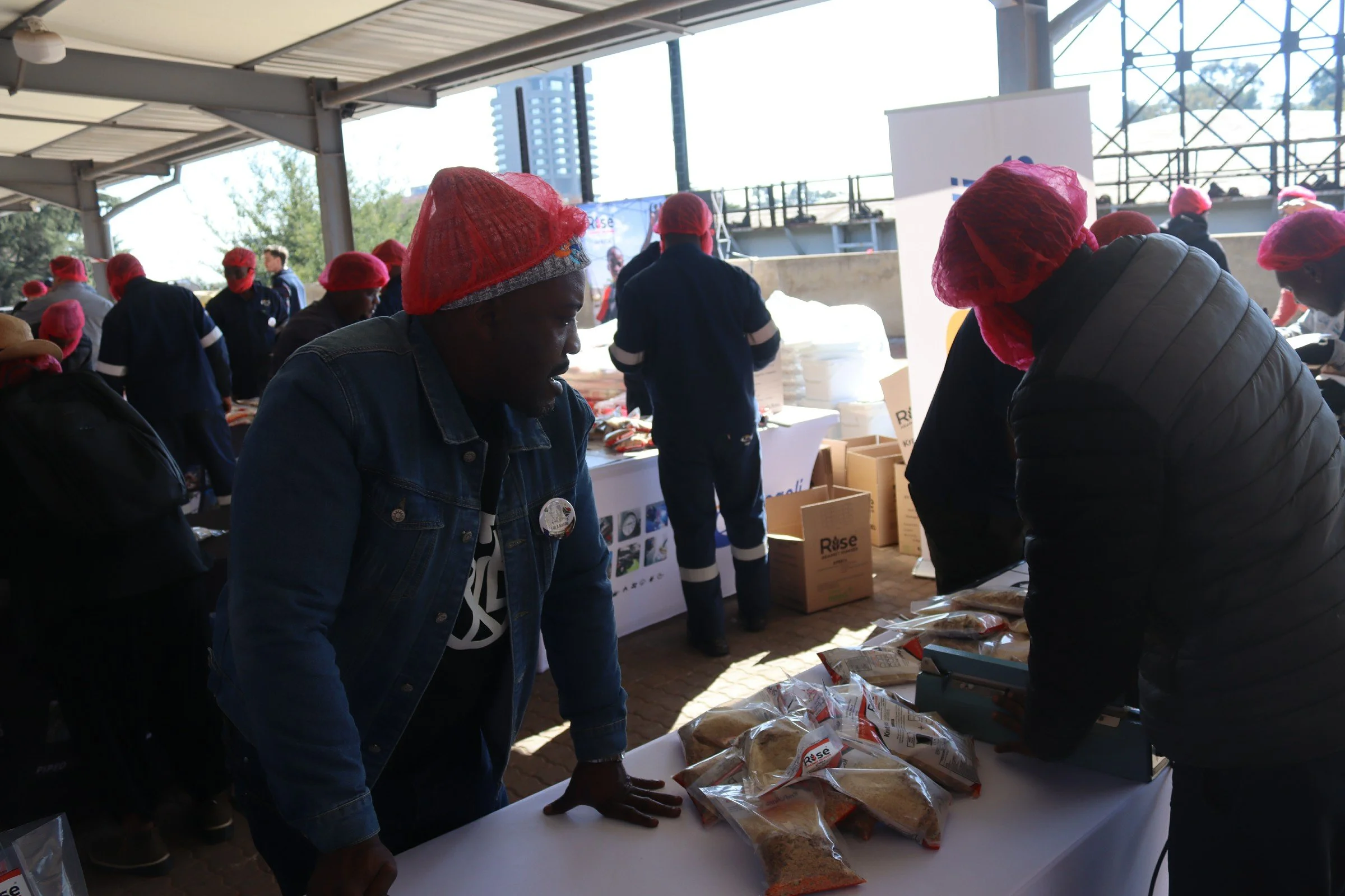 People wearing red hairnets at a table with packaged food items at an outdoor event under a covered area.