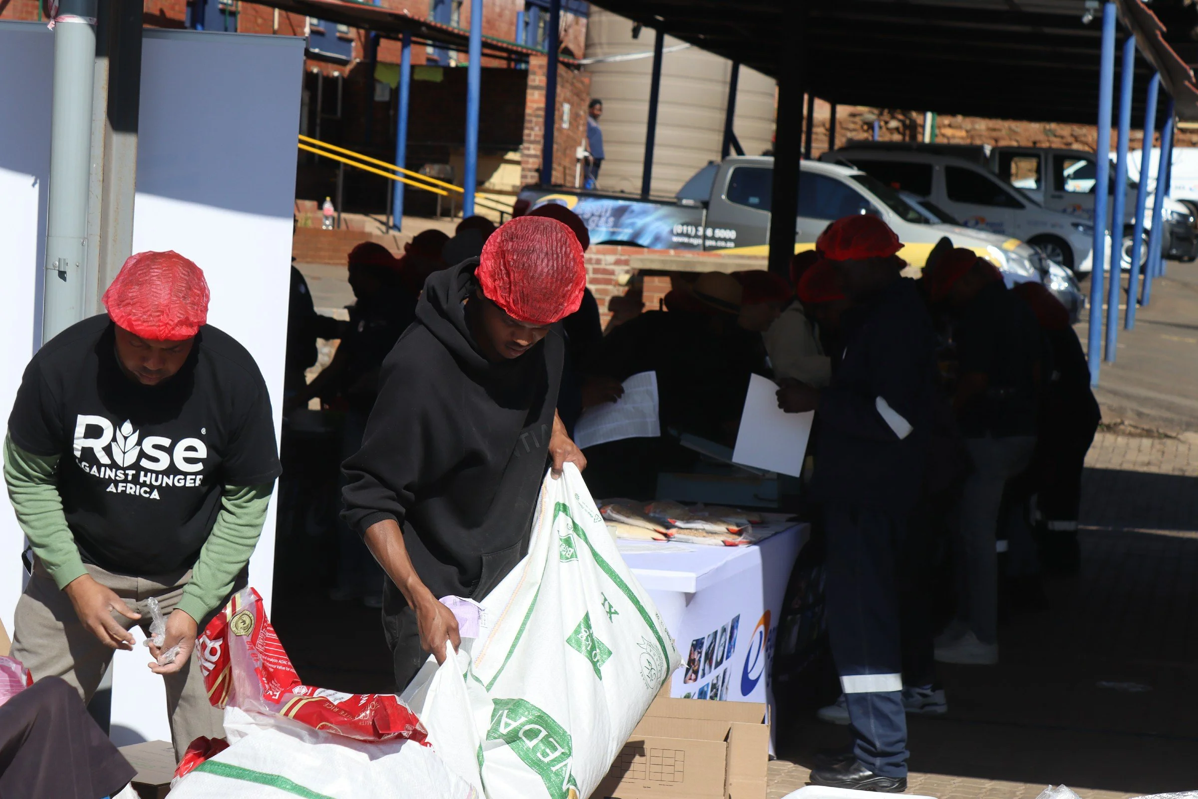 People wearing red hairnets are packing food donations at an outdoor event, with cars and a brick building in the background.