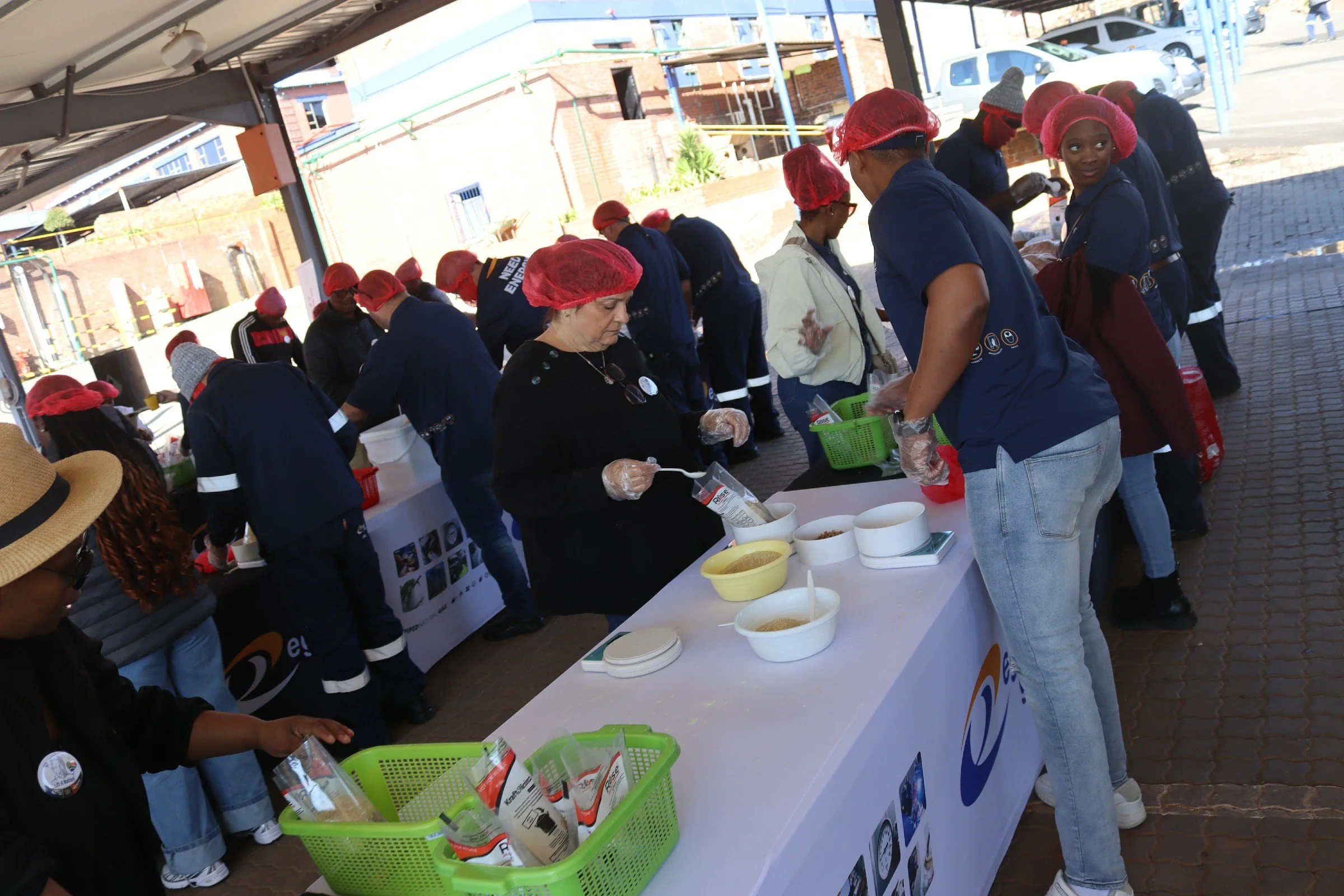 People wearing red hair nets and gloves, standing behind a table with bowls of food, at an outdoor event, possibly a food distribution or community gathering.