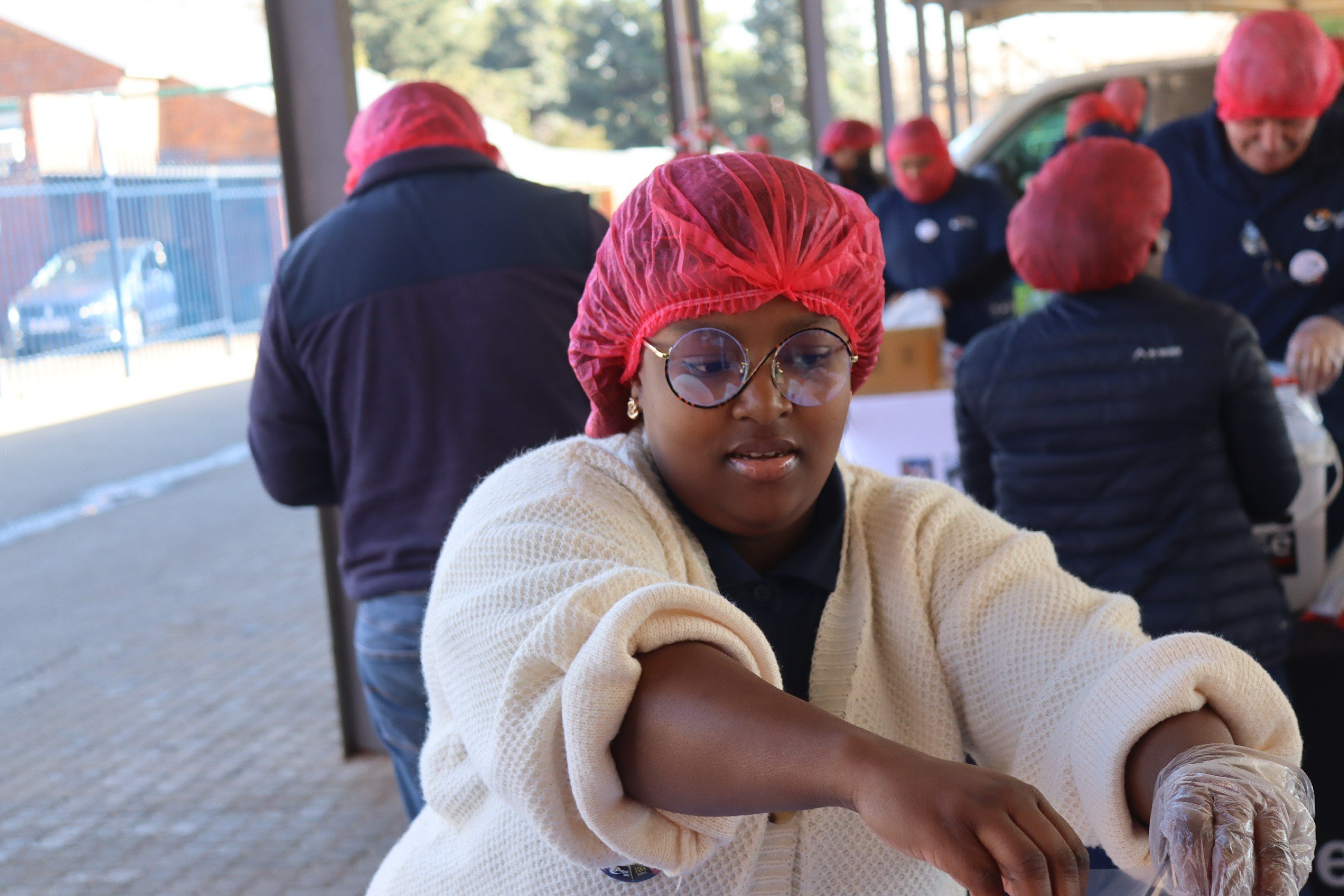 Young girl with glasses and a red hairnet participating in a food packaging event, surrounded by other people also wearing red hairnets, in an outdoor sheltered area.