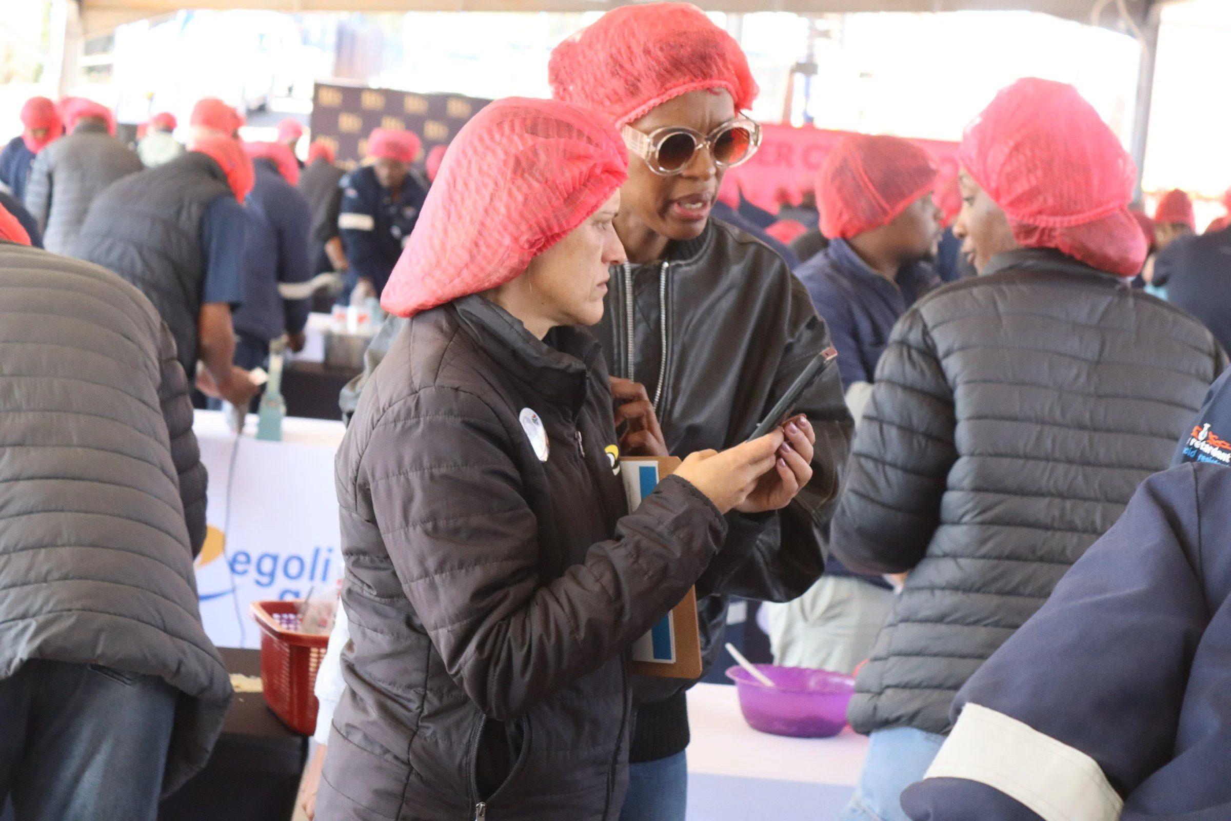 A group of people at an indoor event, many wearing pink hairnets, and one woman in the foreground wearing sunglasses and using her phone.