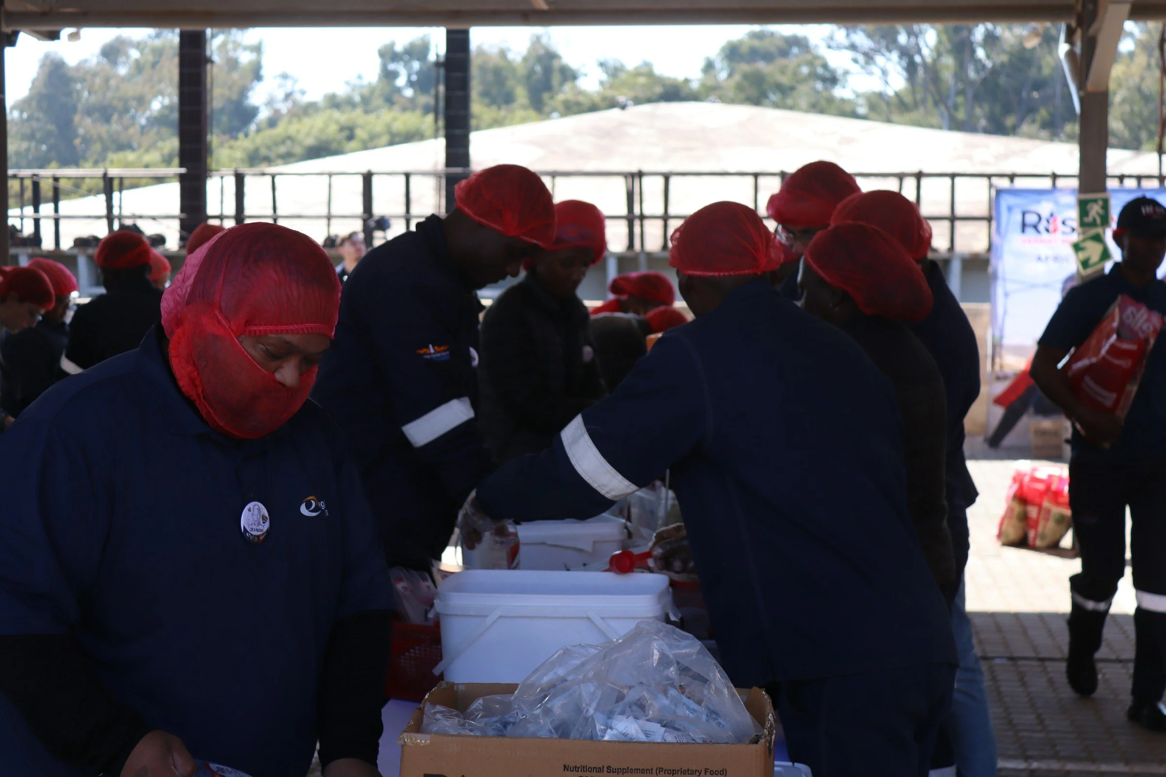 People wearing red hairnets and dark uniforms standing behind tables and packing food in boxes during a community service event outdoors.