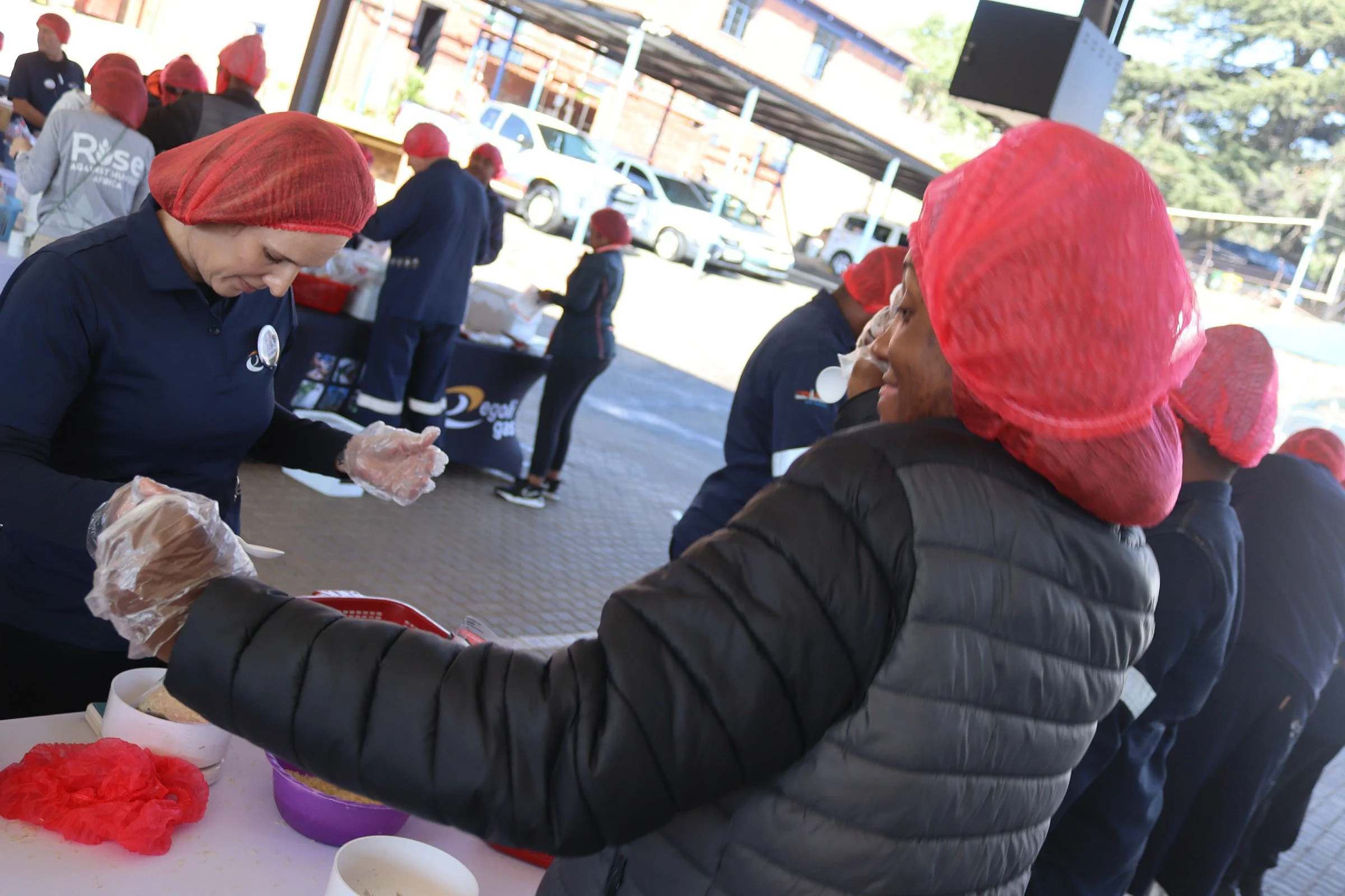 People wearing red hairnets serving food at an outdoor event under a canopy, with cars and trees in the background.