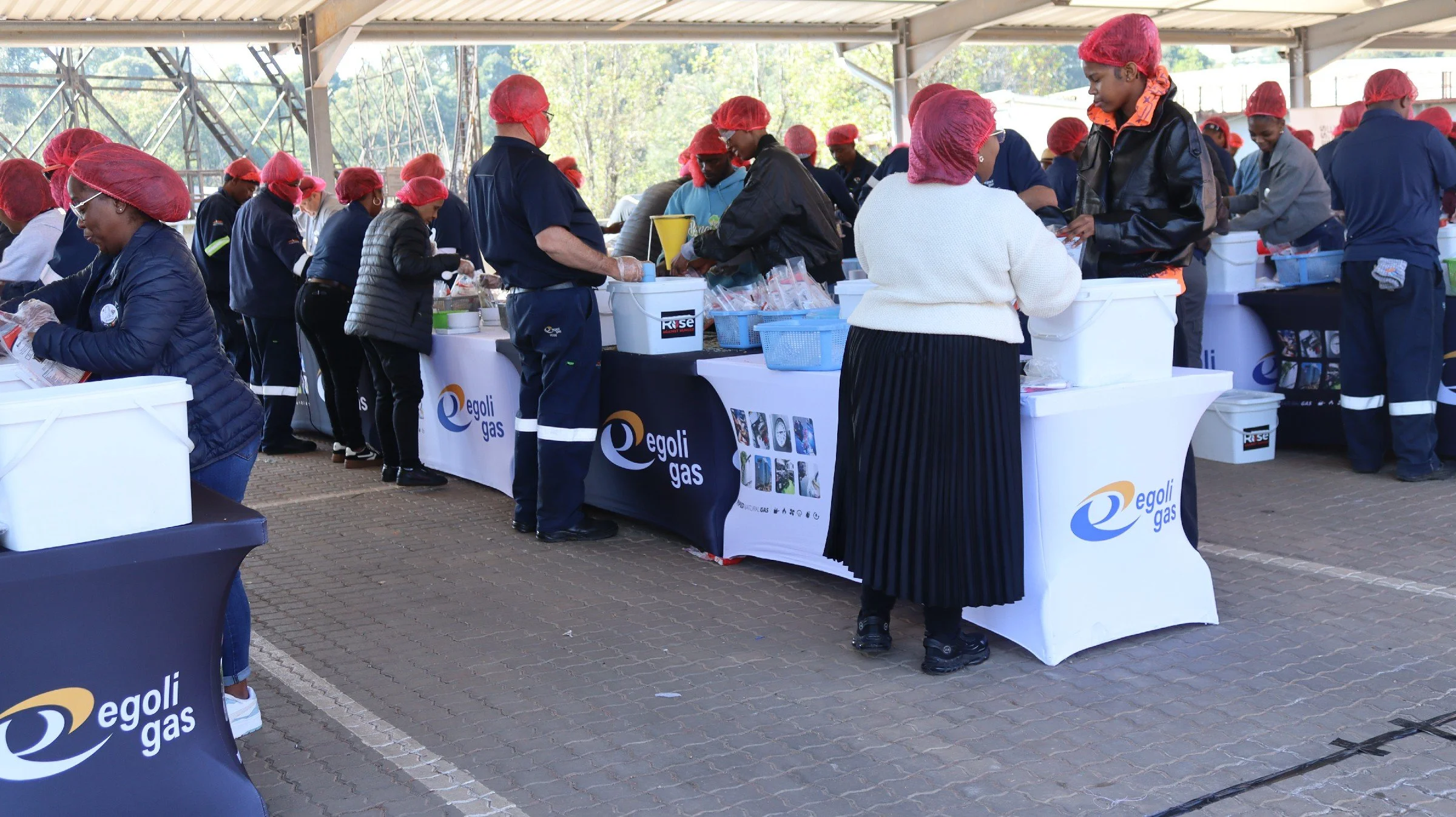 People working at a booth with 'Egol Gas' branding, wearing red hairnets and gloves, serving food or drinks under a large outdoor shelter.
