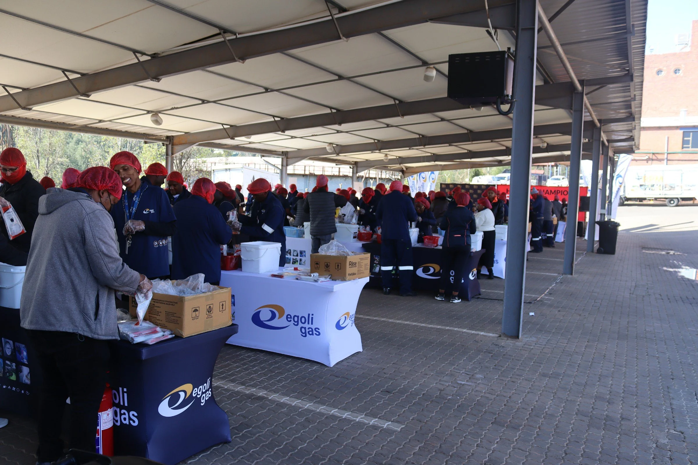 People wearing red hair covers and gloves working at tables with Egoli Gas banners under a covered outdoor area, possibly preparing or serving food.