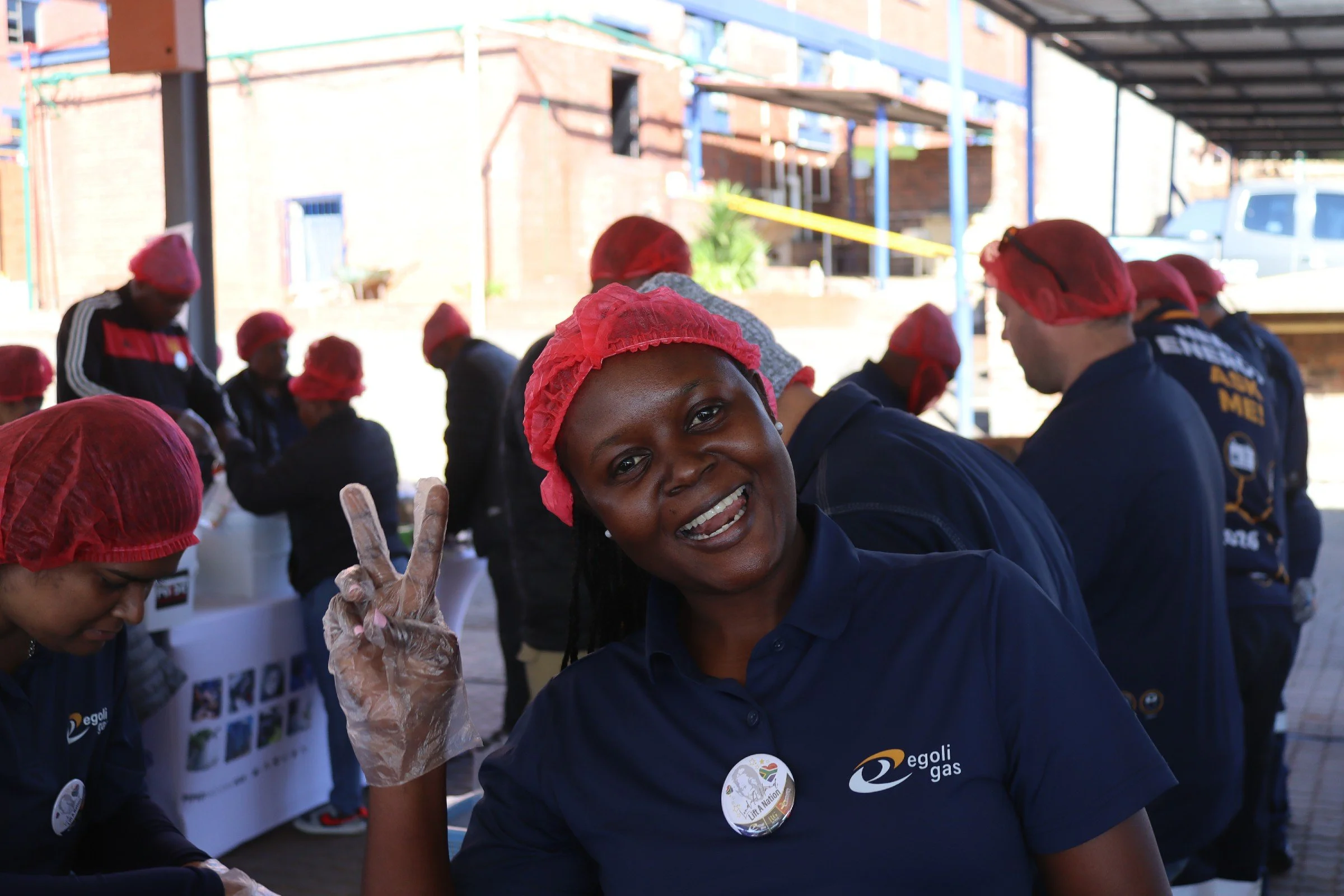 A woman smiling and making a peace sign with her fingers at a gathering of people wearing red hair caps and navy blue shirts, with some standing behind tables with posters in an outdoor setting with buildings in the background.