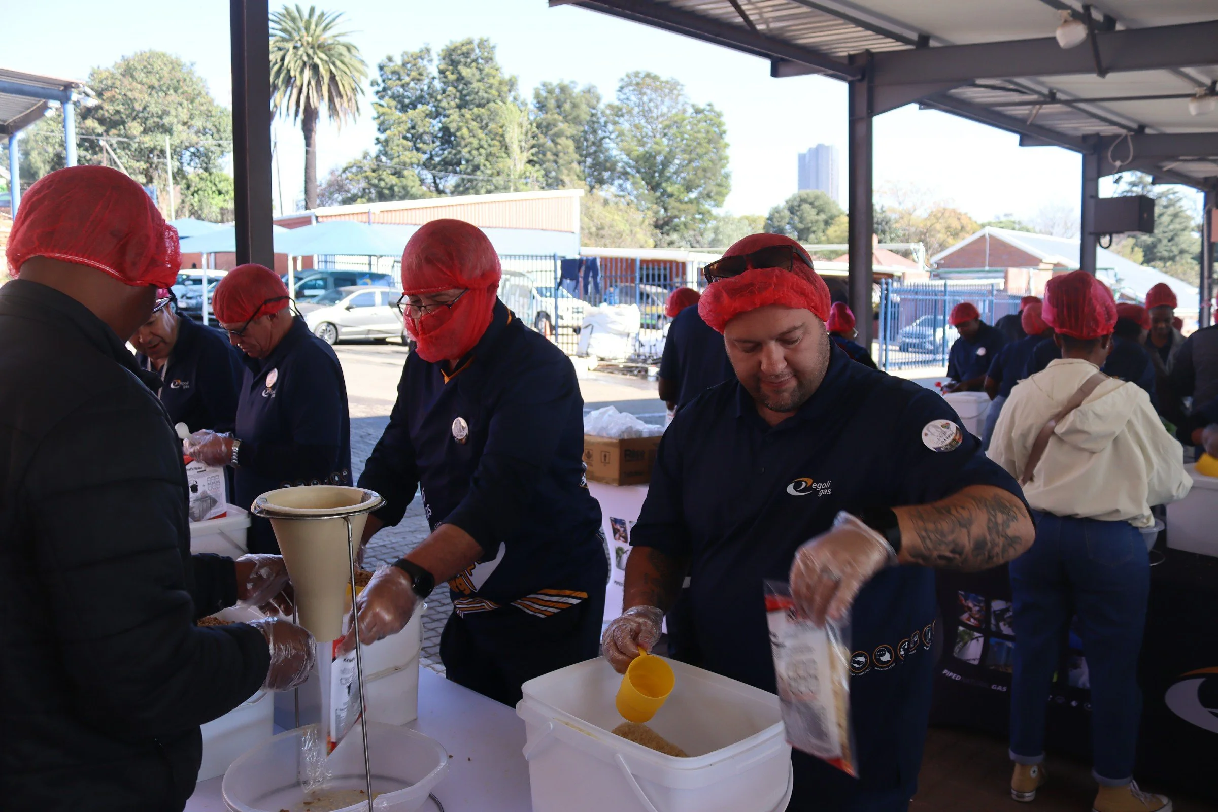 People wearing red hairnets and gloves preparing food at an outdoor event under a covered area, with parking lots, trees, and buildings visible in the background.