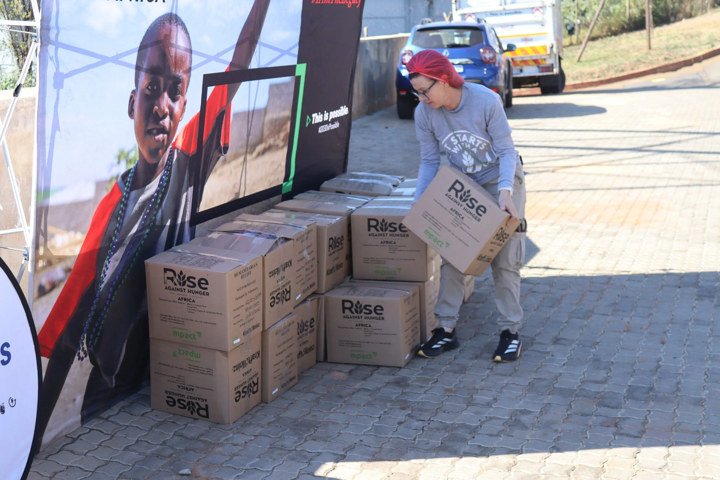 A person wearing glasses, a red headscarf, and a gray shirt with the phrase 'Starts With' is stacking cardboard boxes labeled 'Rise Against Hunger' on a sidewalk next to a large banner. The banner features a portrait of a child and the text 'This is 
