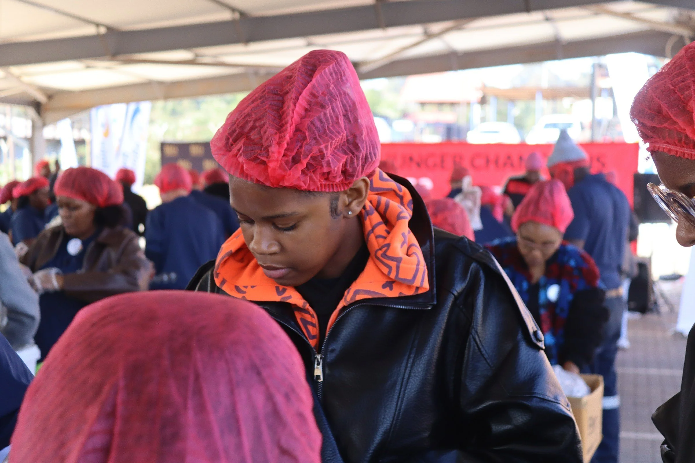A group of people wearing red hairnets gathered under a tent. One person in the foreground is wearing a black leather jacket with an orange patterned scarf.