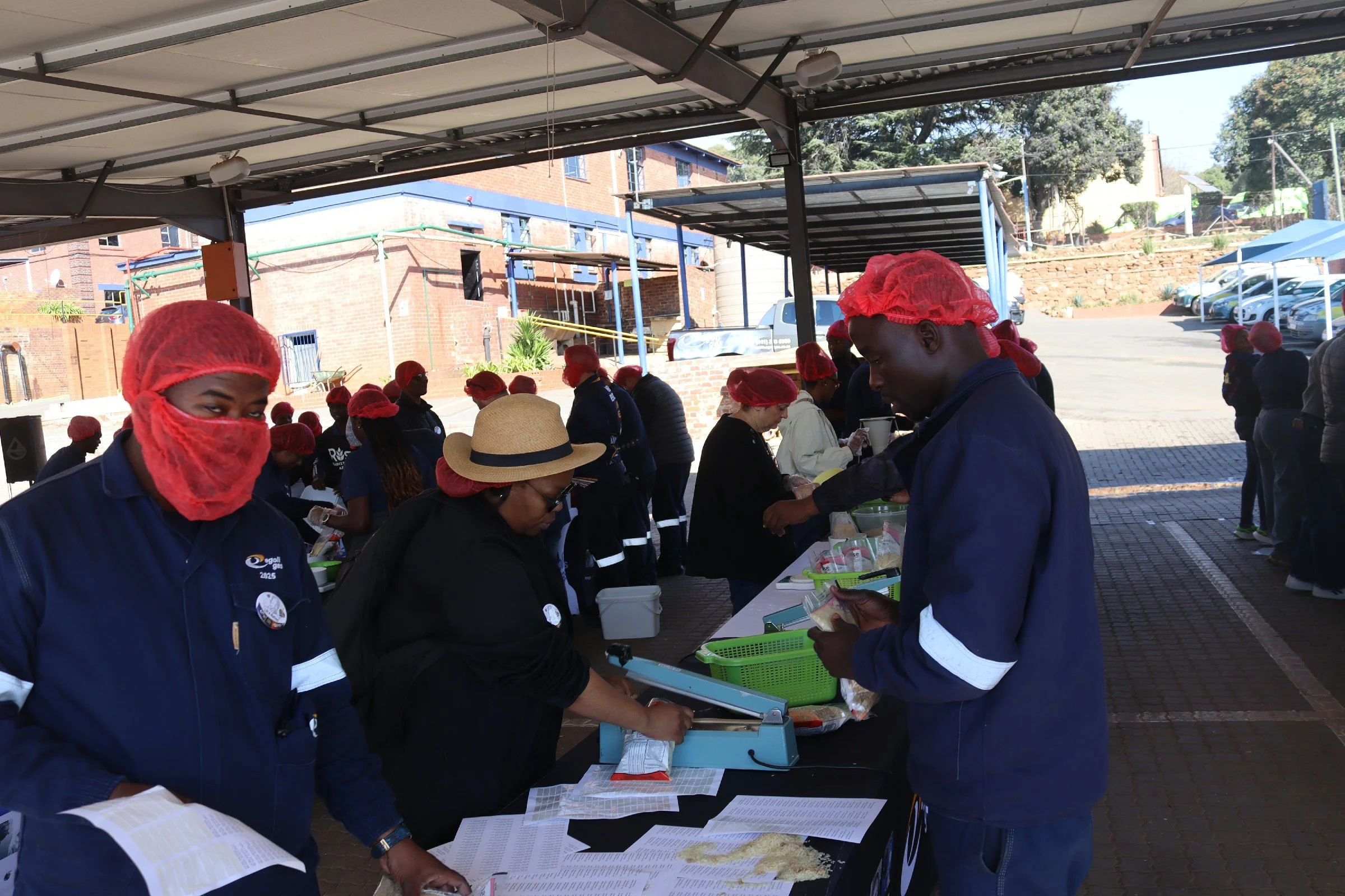 A group of people wearing red hairnets and dark uniforms gathered under a shaded outdoor area, working at a long table with documents and supplies, with a brick building and parking lot in the background.