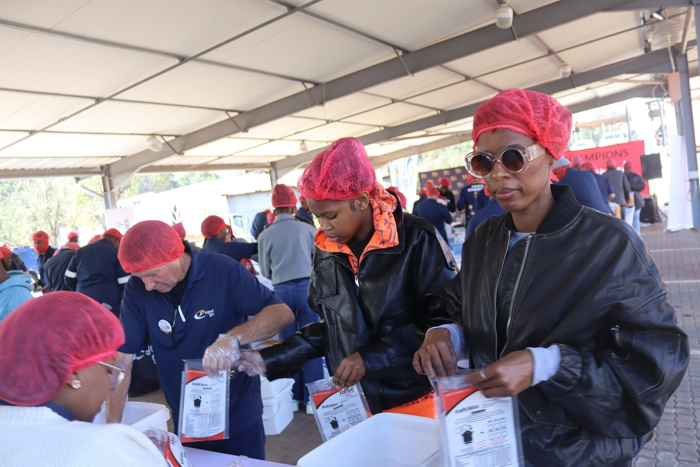 Several people under a large tent, many wearing red hairnets and gloves, participating in what appears to be a community event or charitable activity.
