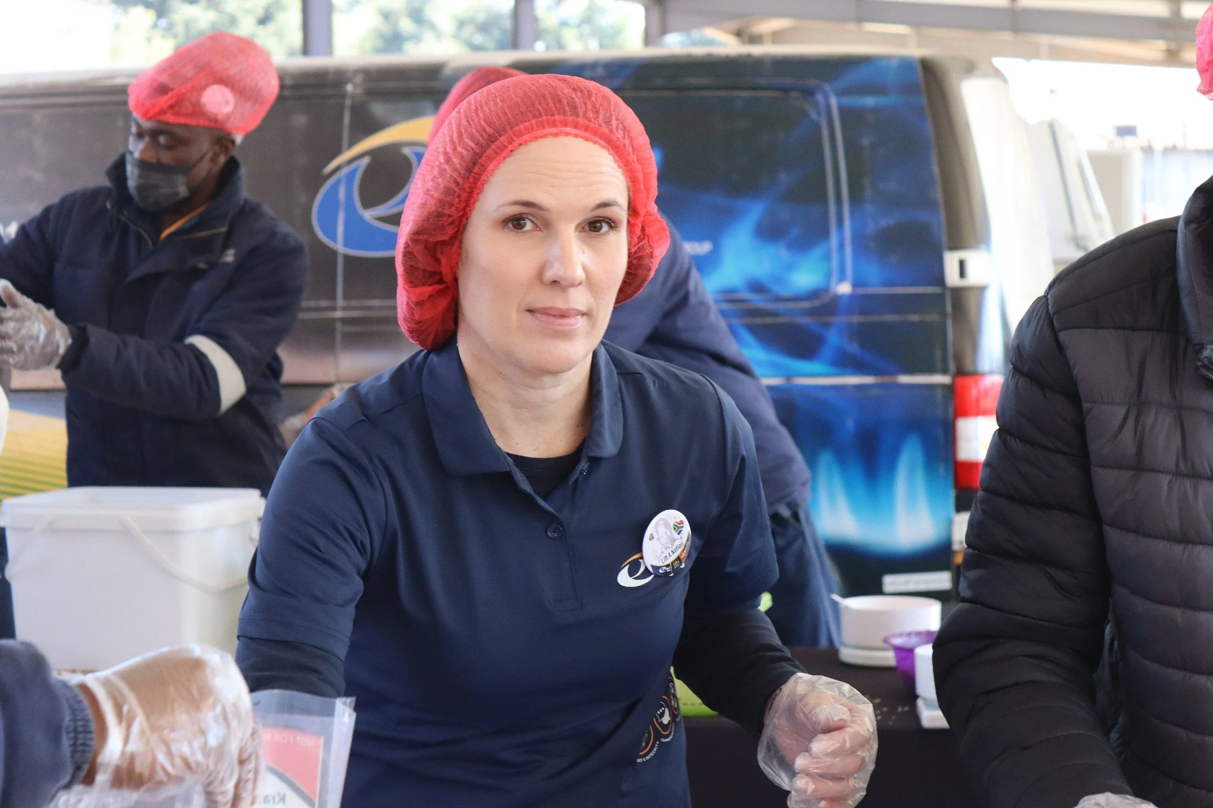 A woman wearing a red hairnet and a dark blue shirt with a badge is participating in a food distribution event, standing near a table with food items, with people working in the background under a covered outdoor setting.