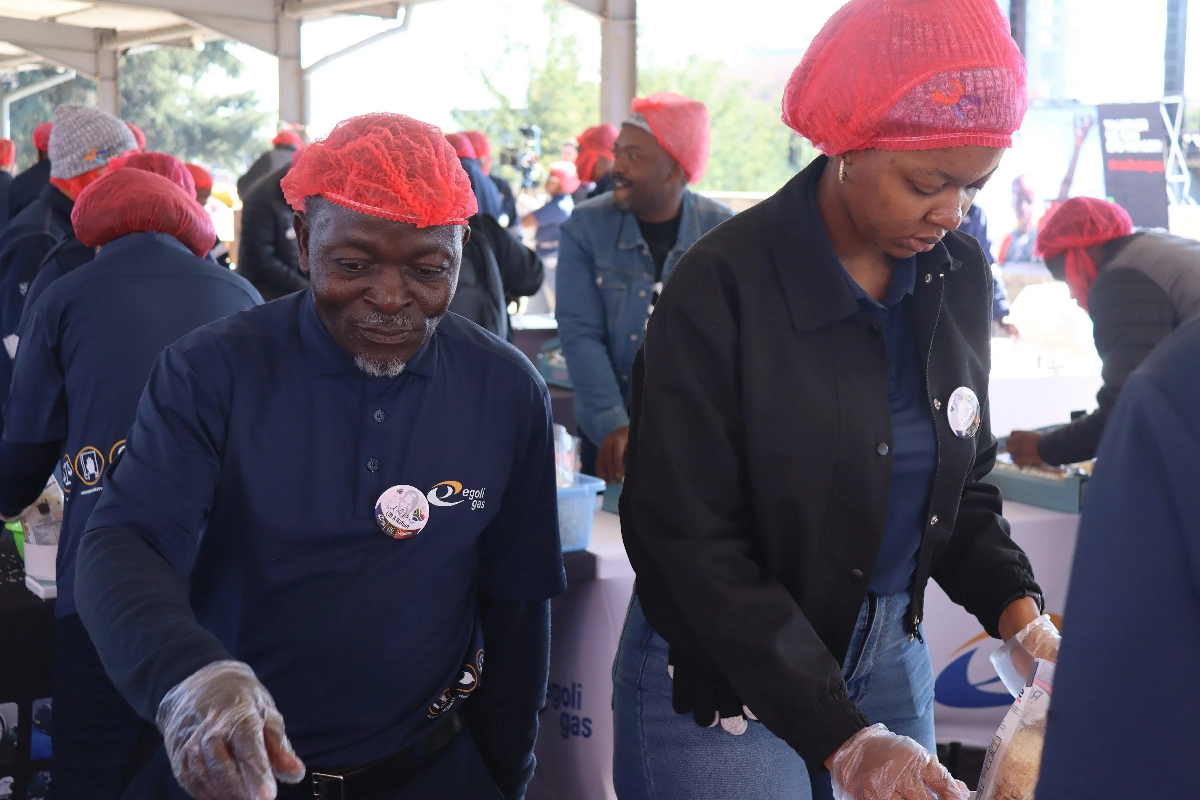 A group of people, mostly women, wearing red hairnets and gloves, participating in a food packaging or distribution event under a tent outdoors.