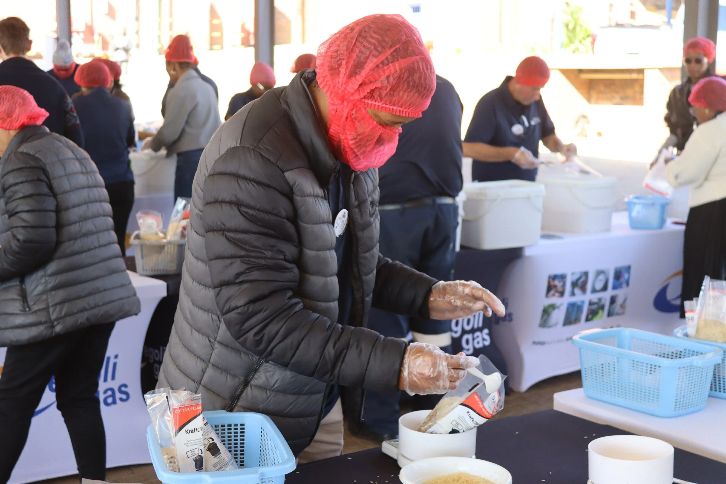 People wearing red hair nets and gloves at a food preparation or packaging event. They are working at tables with various containers and packages.