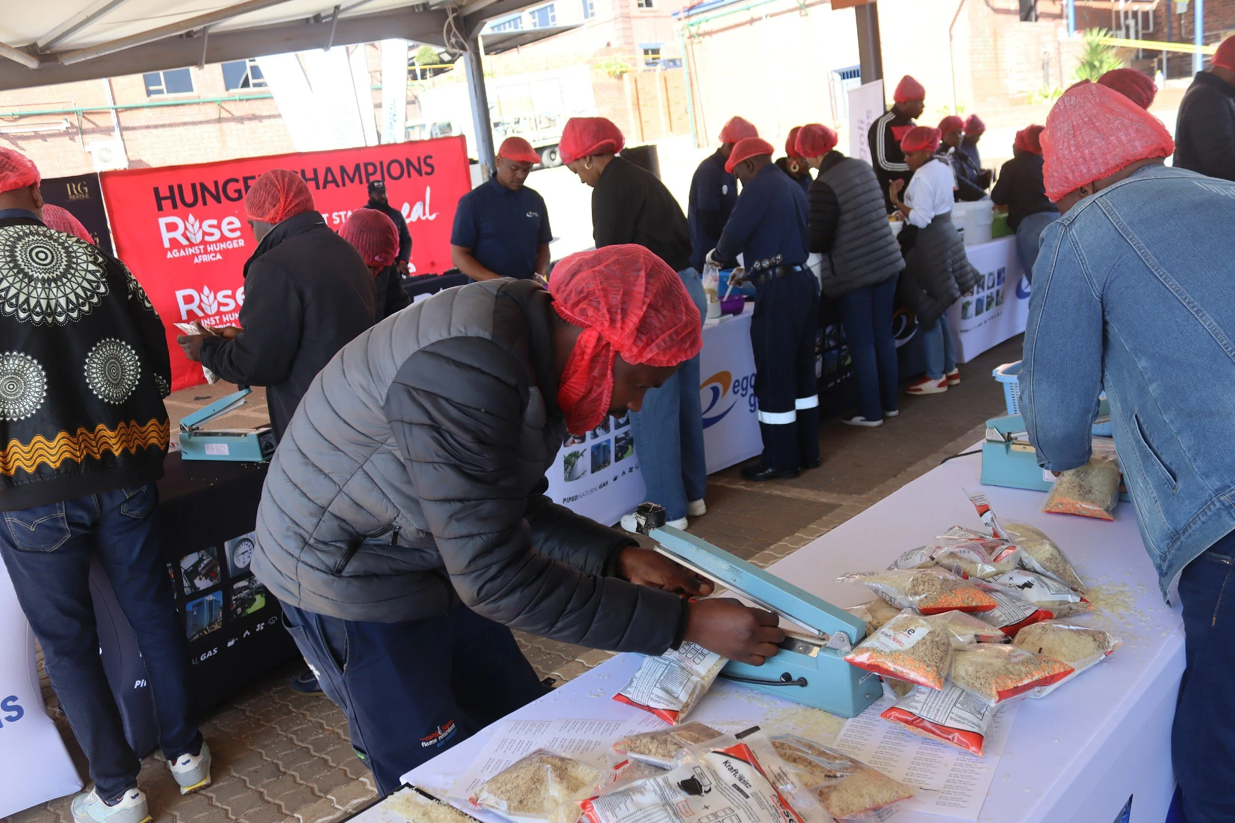 People participating in a food distribution event, wearing red hairnets, standing in line, and selecting food items such as rice and grains from tables.