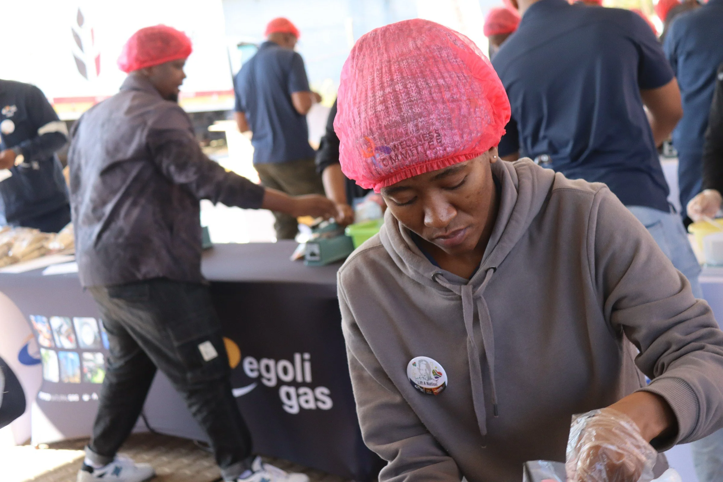 People wearing red caps and working at a table during an event, some are focused on tasks, with banners and digital screens displaying text and logos in the background.