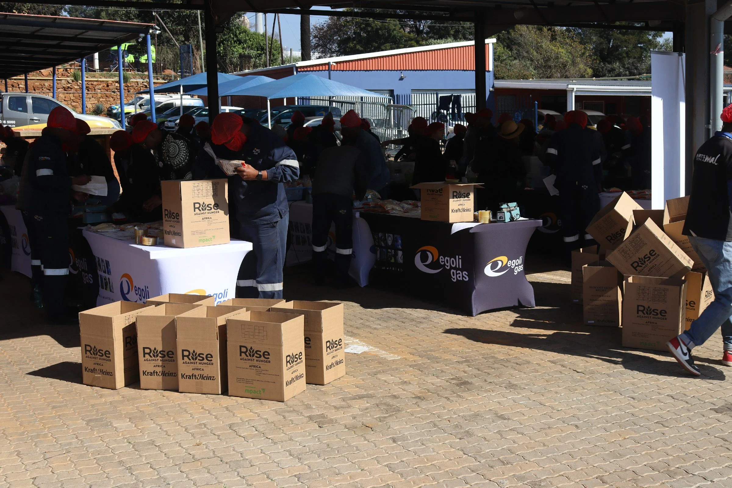 People participating in a charity event or fundraiser, distributing food and supplies under an outdoor shelter, with boxes of goods labeled 'Rise Against Hunger' on the tables and ground, and some individuals reading or handling papers.