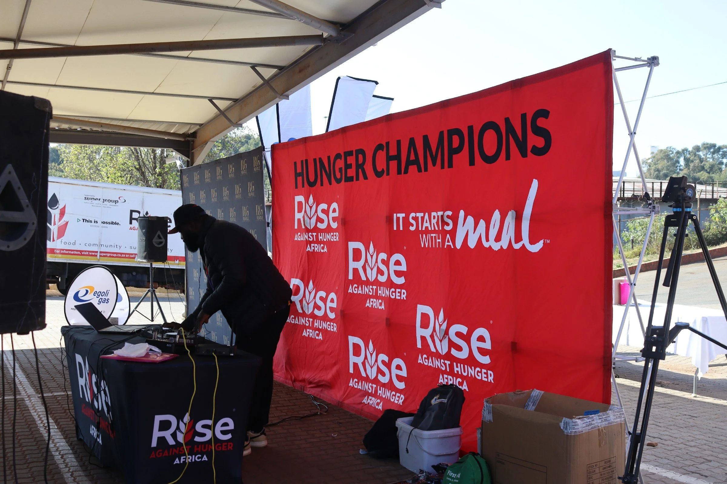 Event setup with banners and a DJ station at a Rise Against Hunger Africa charity event, under a white canopy with trees and a bridge in the background.
