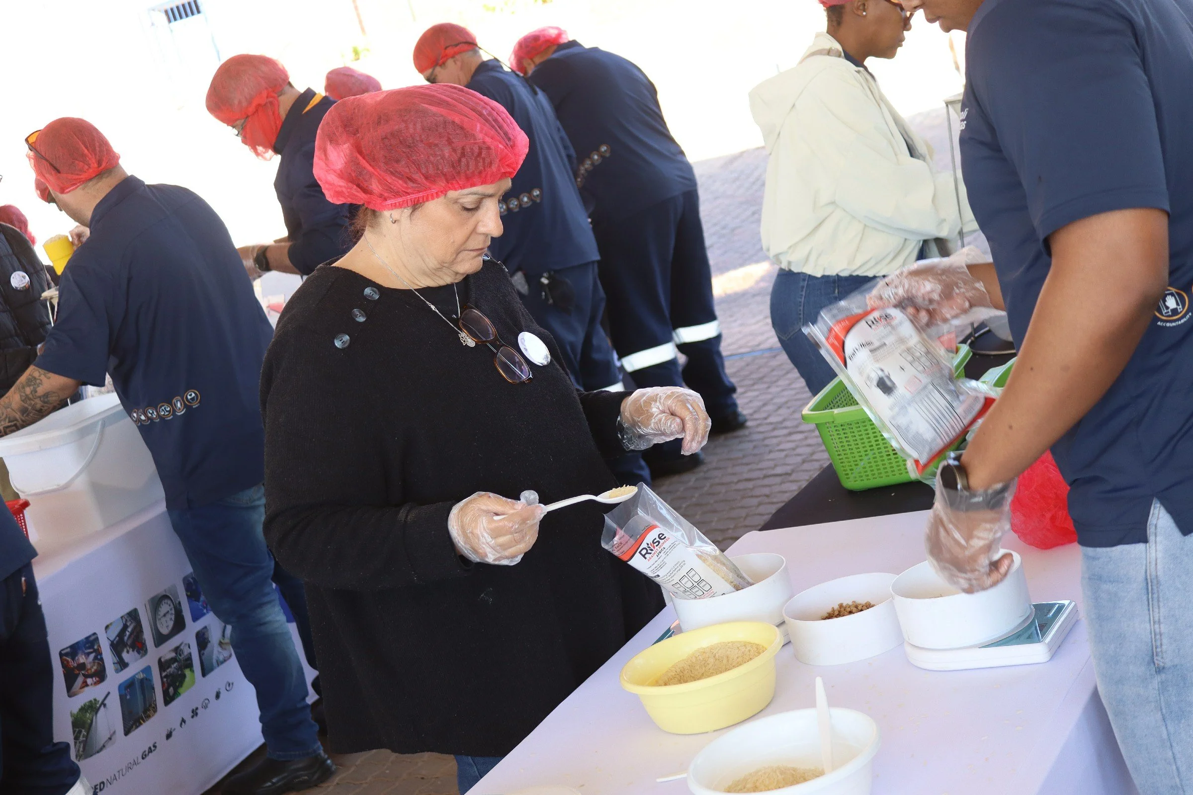 People in hairnets and gloves preparing food at a table with various ingredients like grains and spices, under an outdoor covered area.