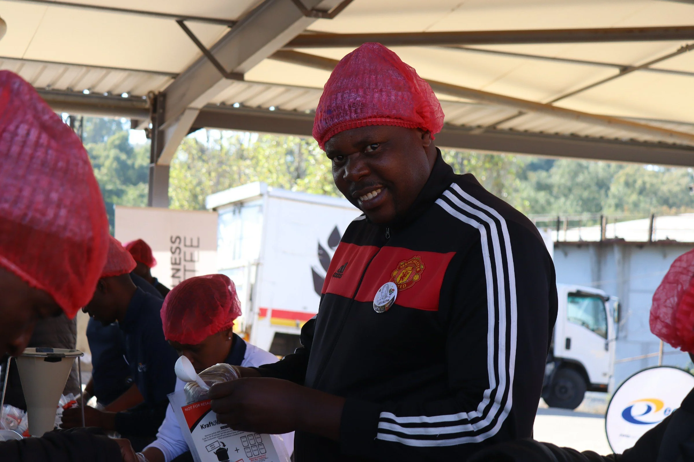 A man wearing a Manchester United jacket and a red hairnet is smiling while participating in an outdoor event under a covered structure, with other people wearing similar hairnets around him.