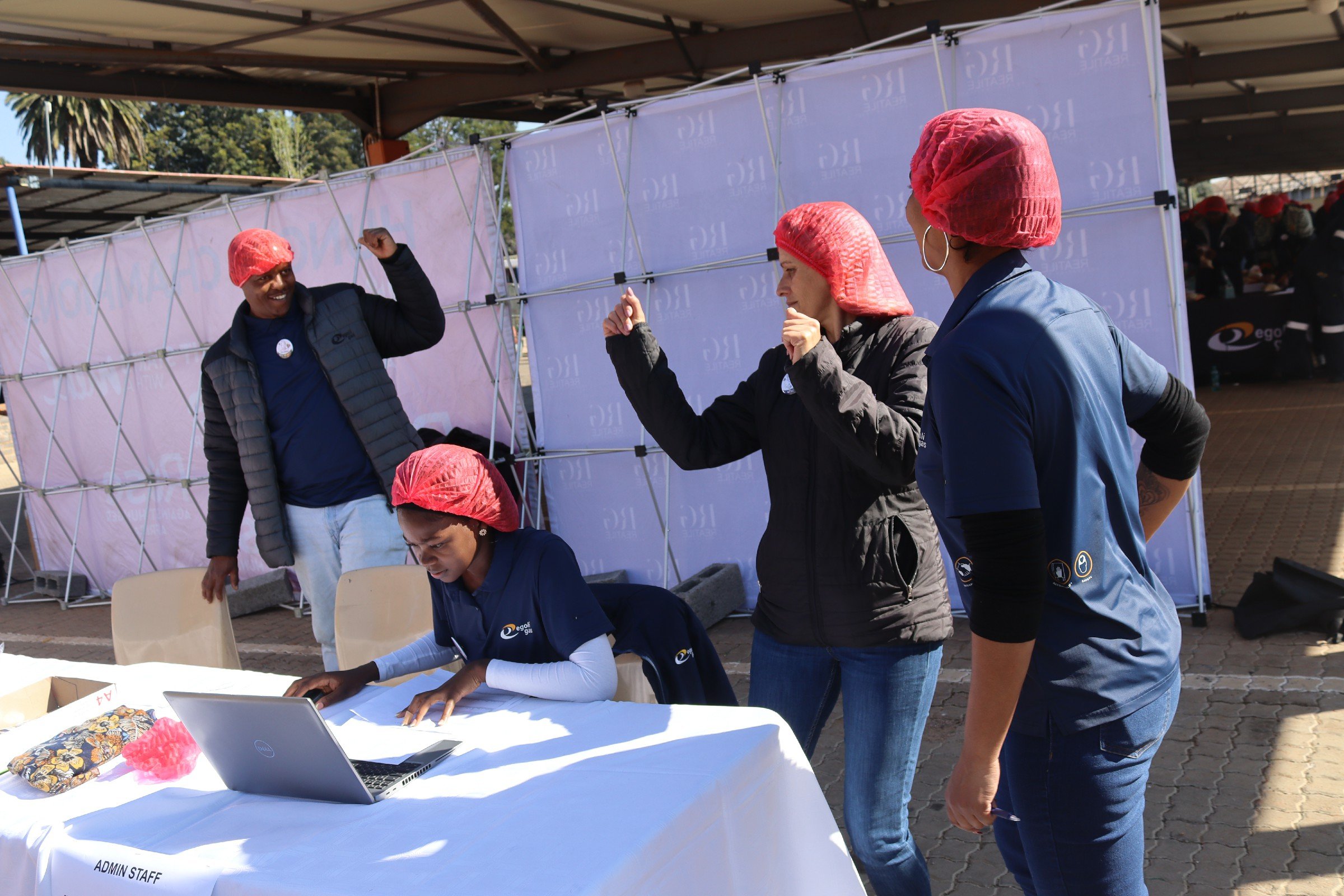People wearing pink hairnets and dark jackets gather around a table with a laptop at an outdoor event, under a canopy with purple banners.