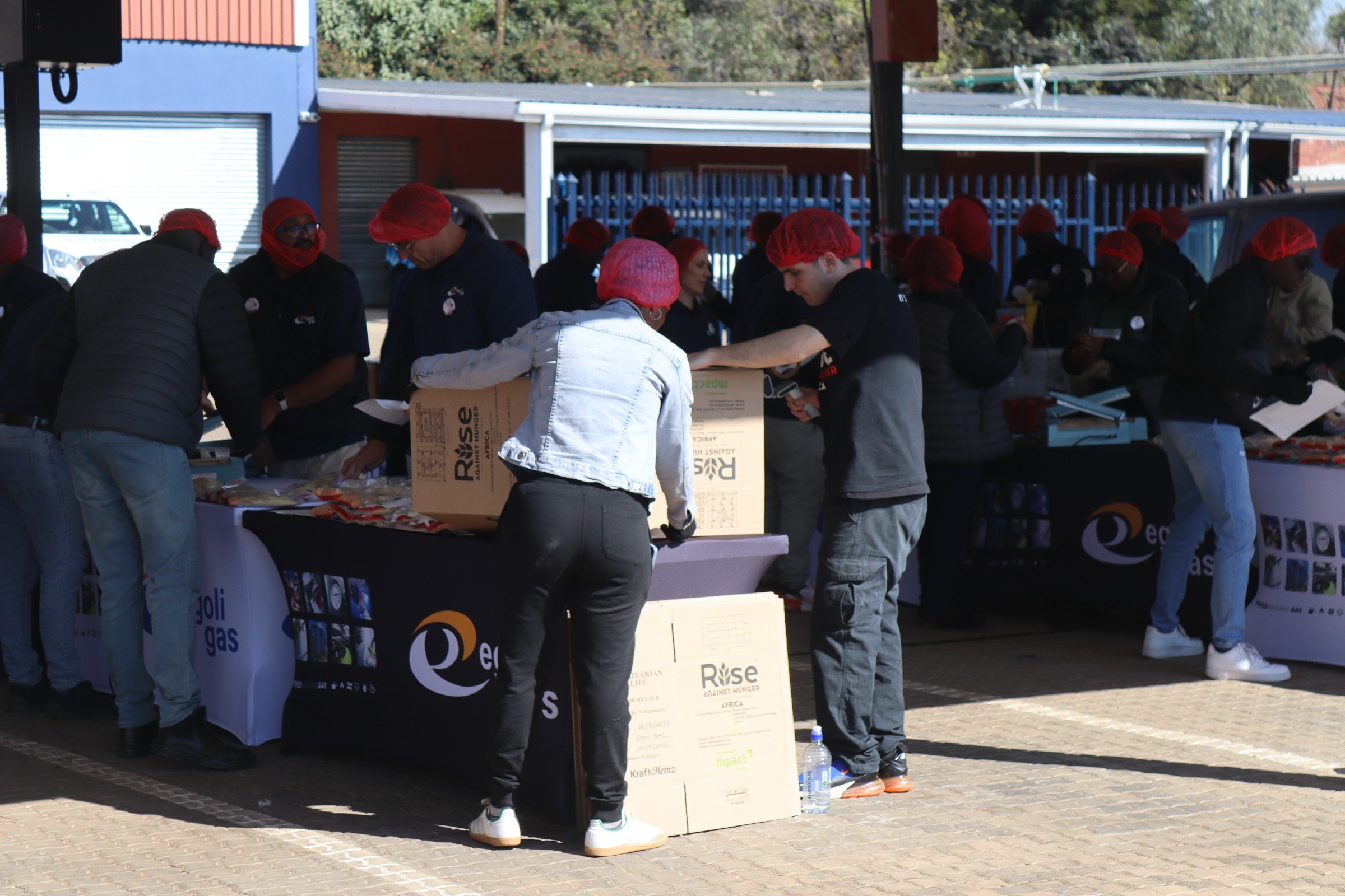People wearing red hairnets and dark clothing at an outdoor charity event, distributing or organizing food under a tent with tables and informational banners.