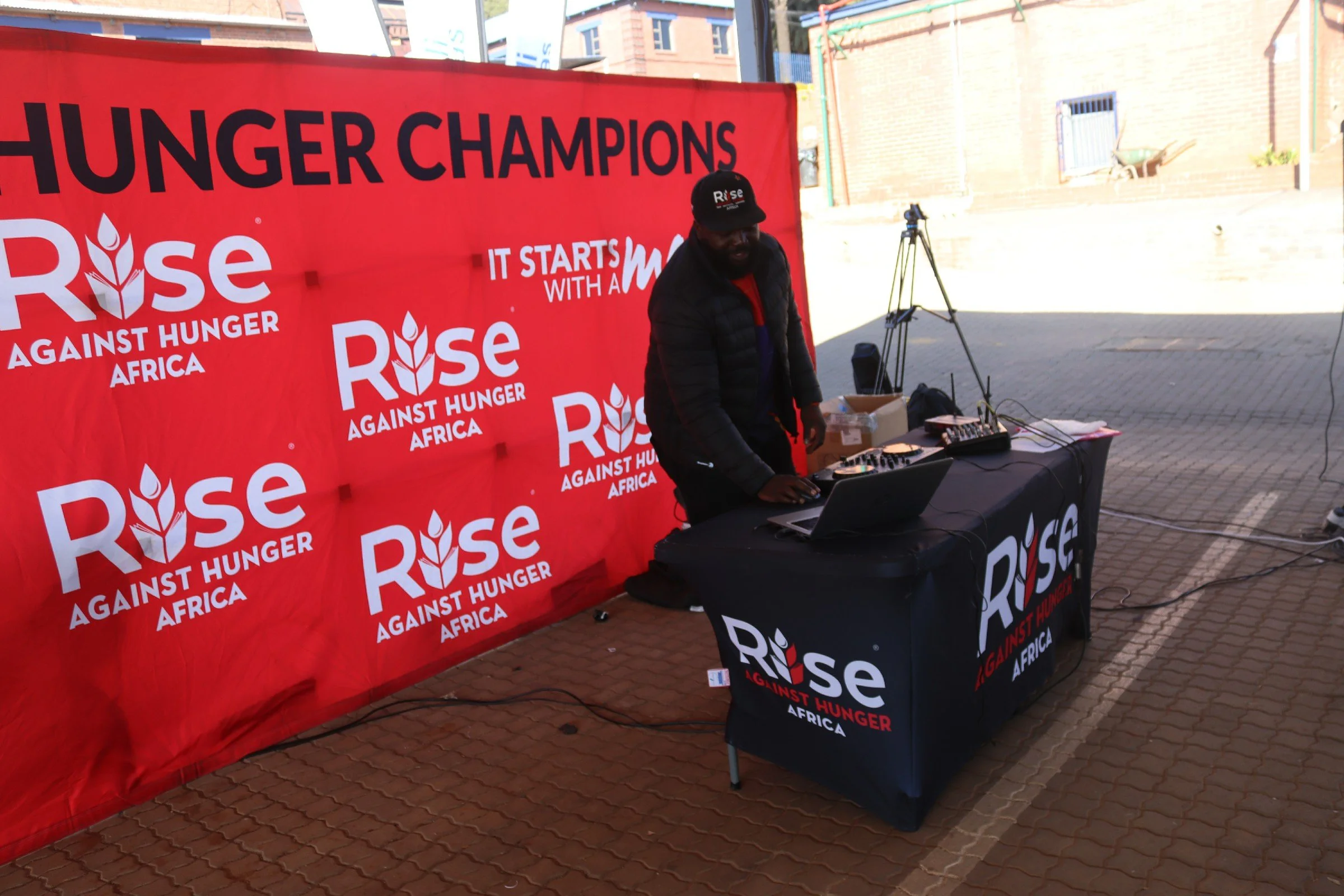 A DJ stands behind a black table with a laptop, DJ equipment, and a Rise Against Hunger Africa banner, at an outdoor event with a red Rise Against Hunger Africa backdrop.