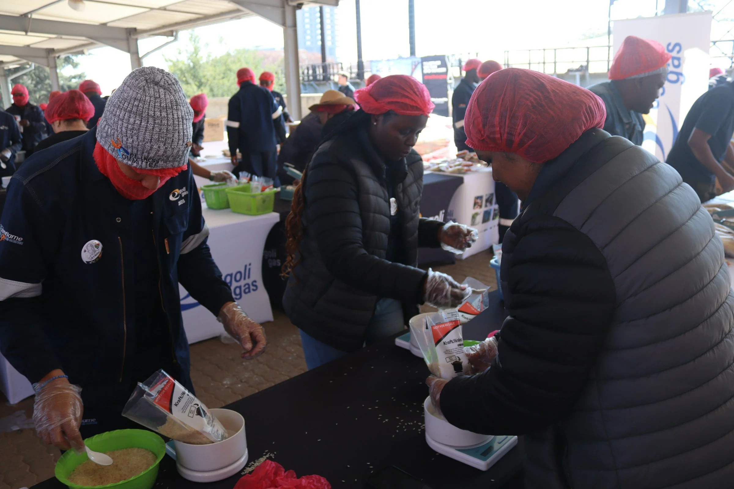 Group of people preparing food at an outdoor event under a covered pavilion, with some wearing red hair covers and gloves.
