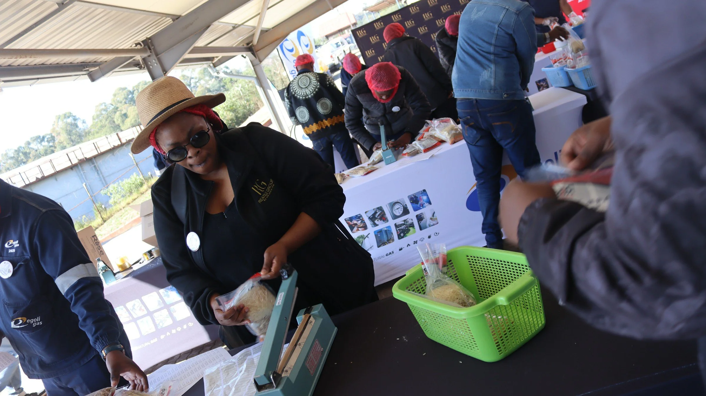 People at an outdoor market or event under a tent, with some wearing red hairnets, handling packages of food or products at a booth with informational posters and banners.