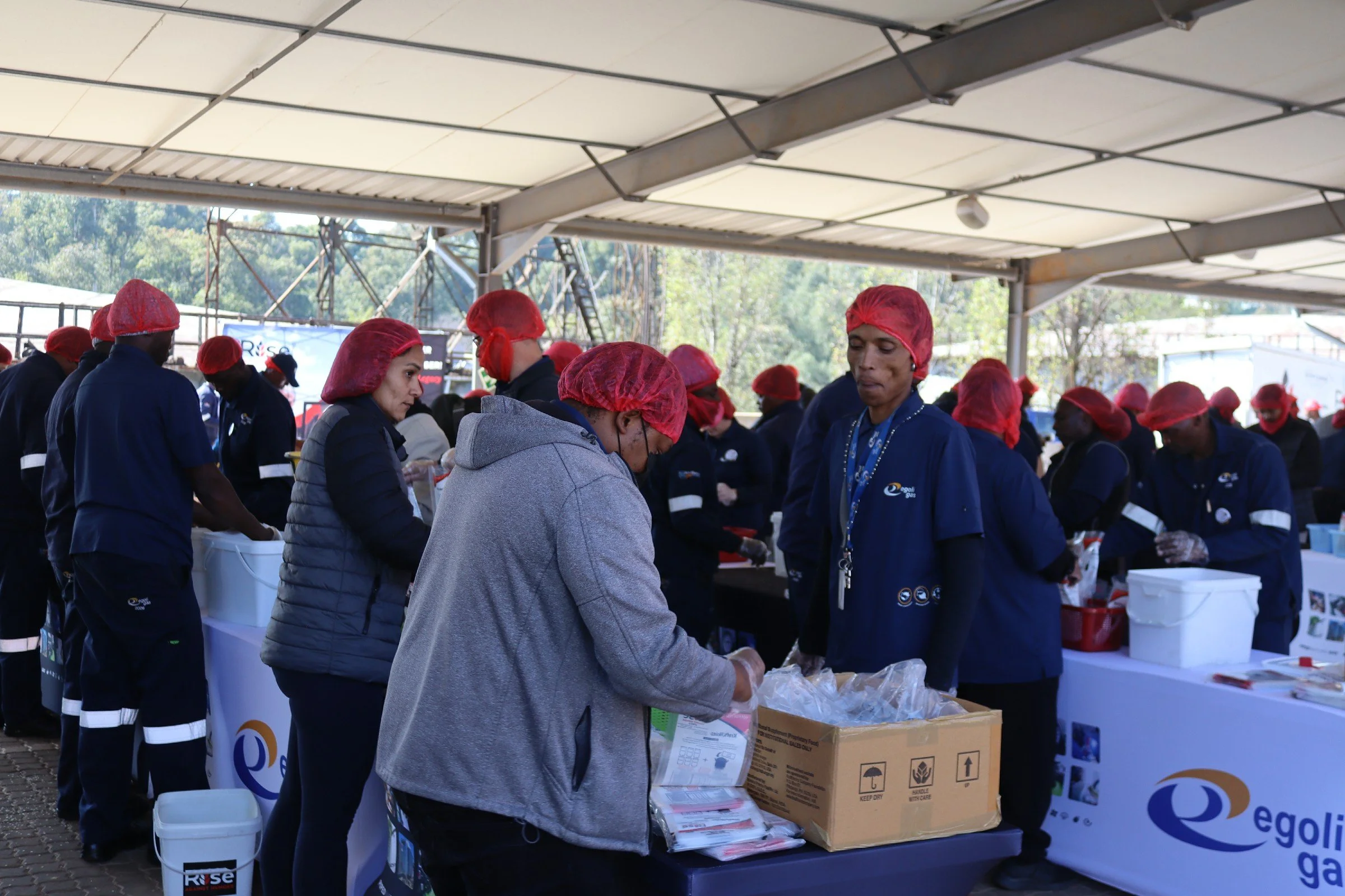 People wearing blue uniforms and red hair covers working at an outdoor event under a tent, packing supplies into boxes, with trees and equipment visible in the background.