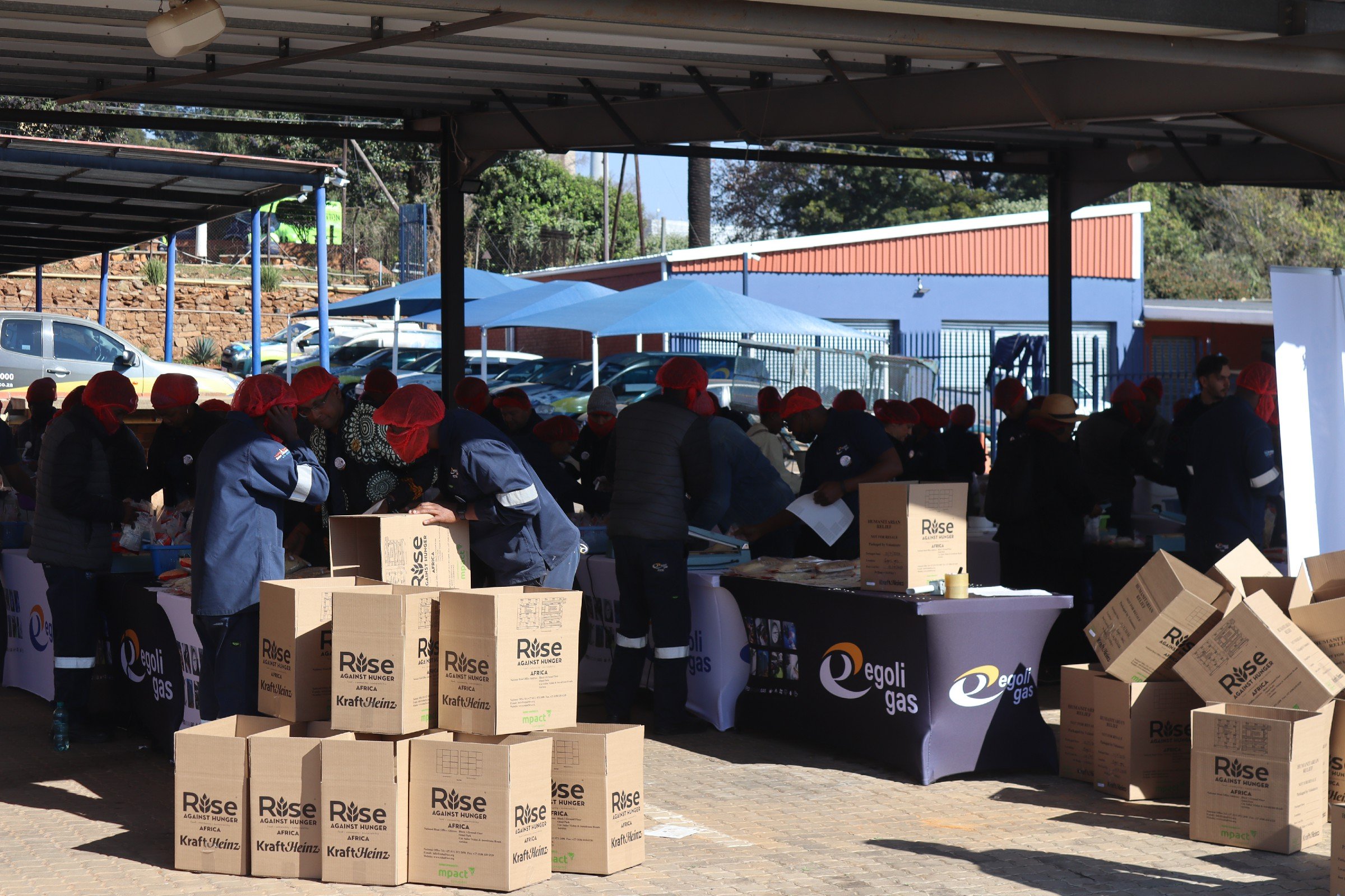 People wearing red head coverings organize boxes with 'Rise Against Hunger' and 'Kraft Heinz' logos at a charity event under a shaded outdoor area, with parked cars and a building in the background.