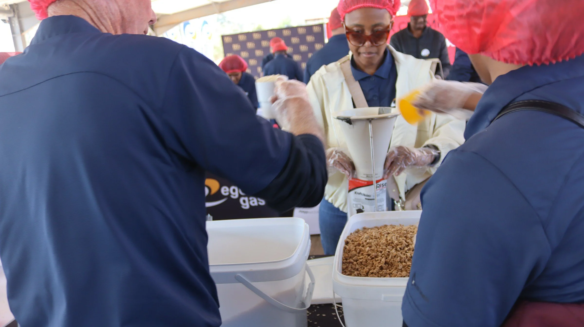 People serving food at a community event with large containers of cooked food and packaging supplies.