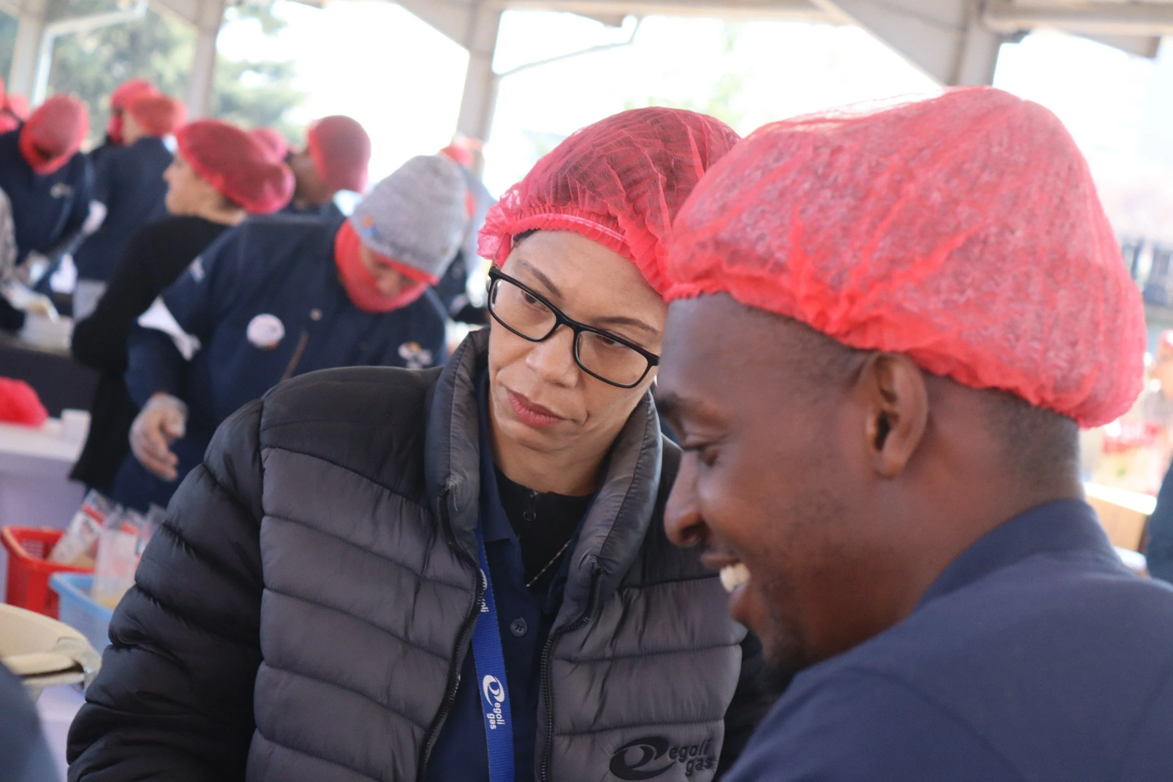 Two women wearing pink hairnets and dark jackets are working together at a table in a busy indoor setting with other people in pink hairnets in the background.