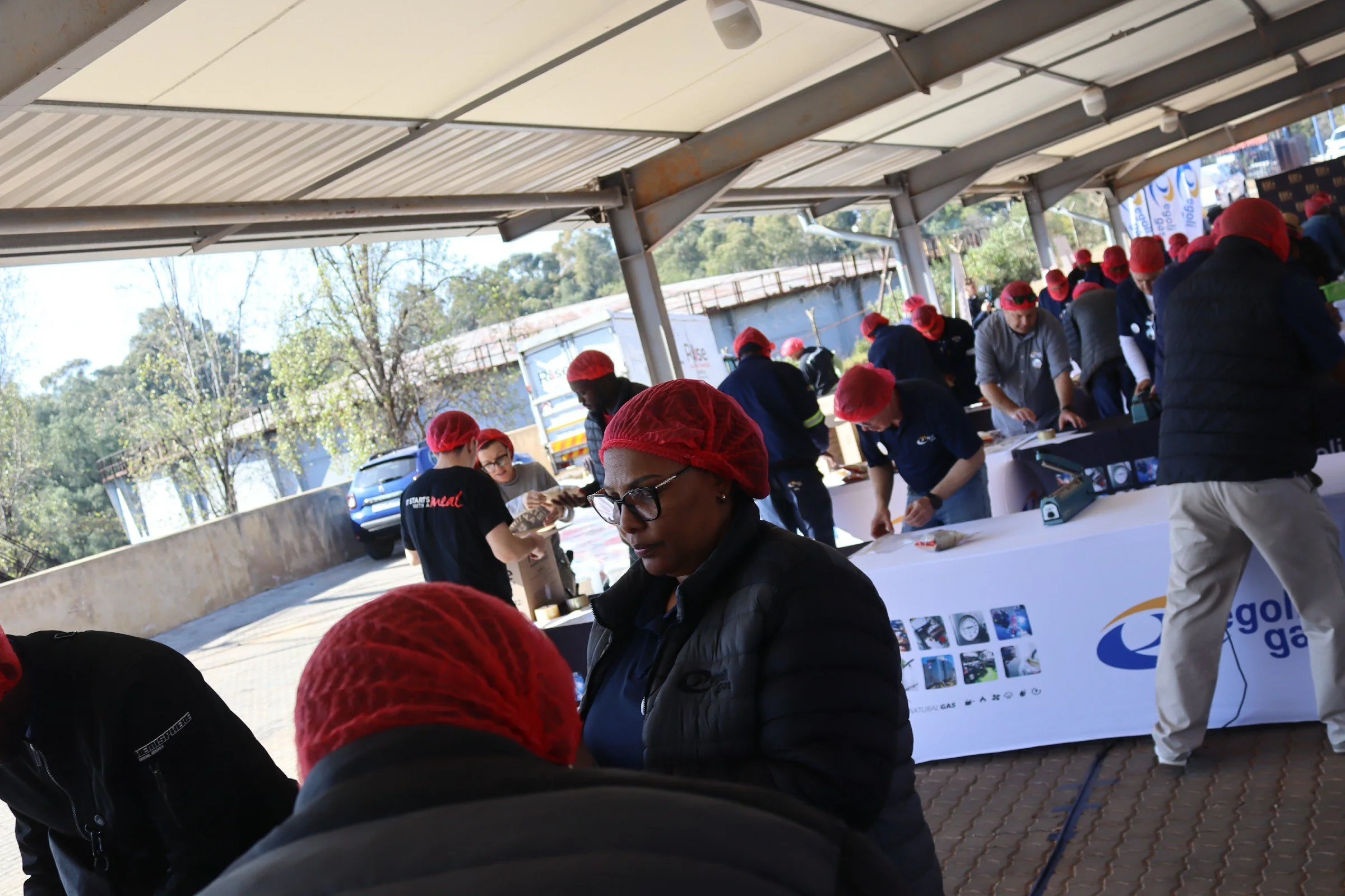 People wearing red hairnets gather around tables at an outdoor event under a metal awning, with banners and trees in the background.