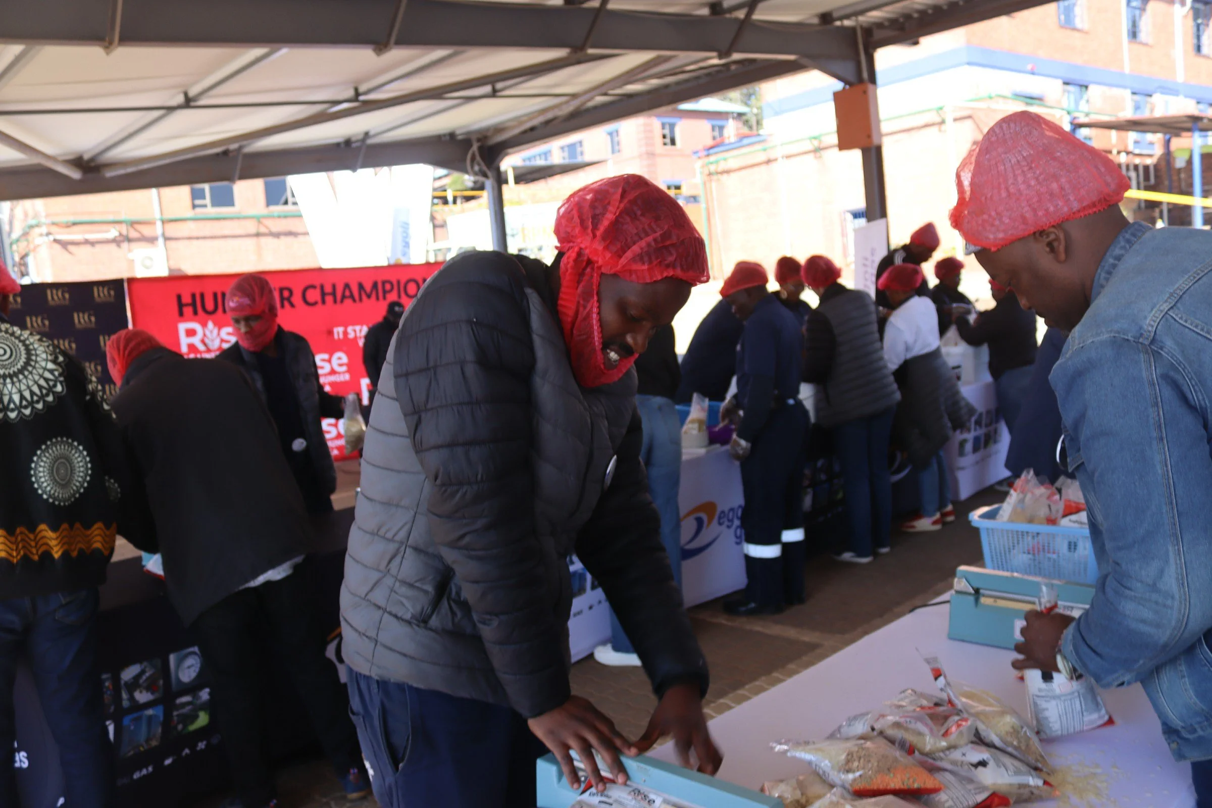 People shopping at an outdoor market, wearing red hairnets, with a red banner in the background reading 'HUI - THE CHAMPION'.