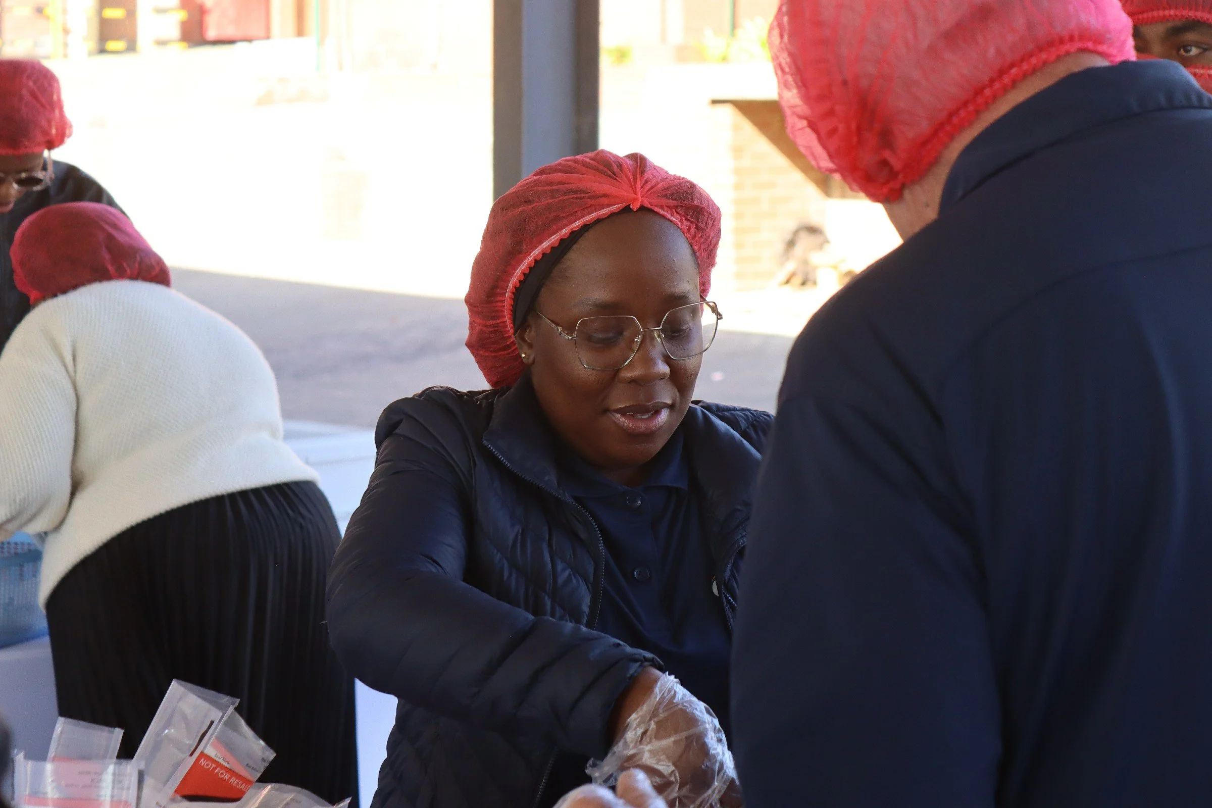 A group of people, mostly women, wearing red hair caps, engaged in a communal activity, possibly at a market or distribution event, with some handling plastic bags.
