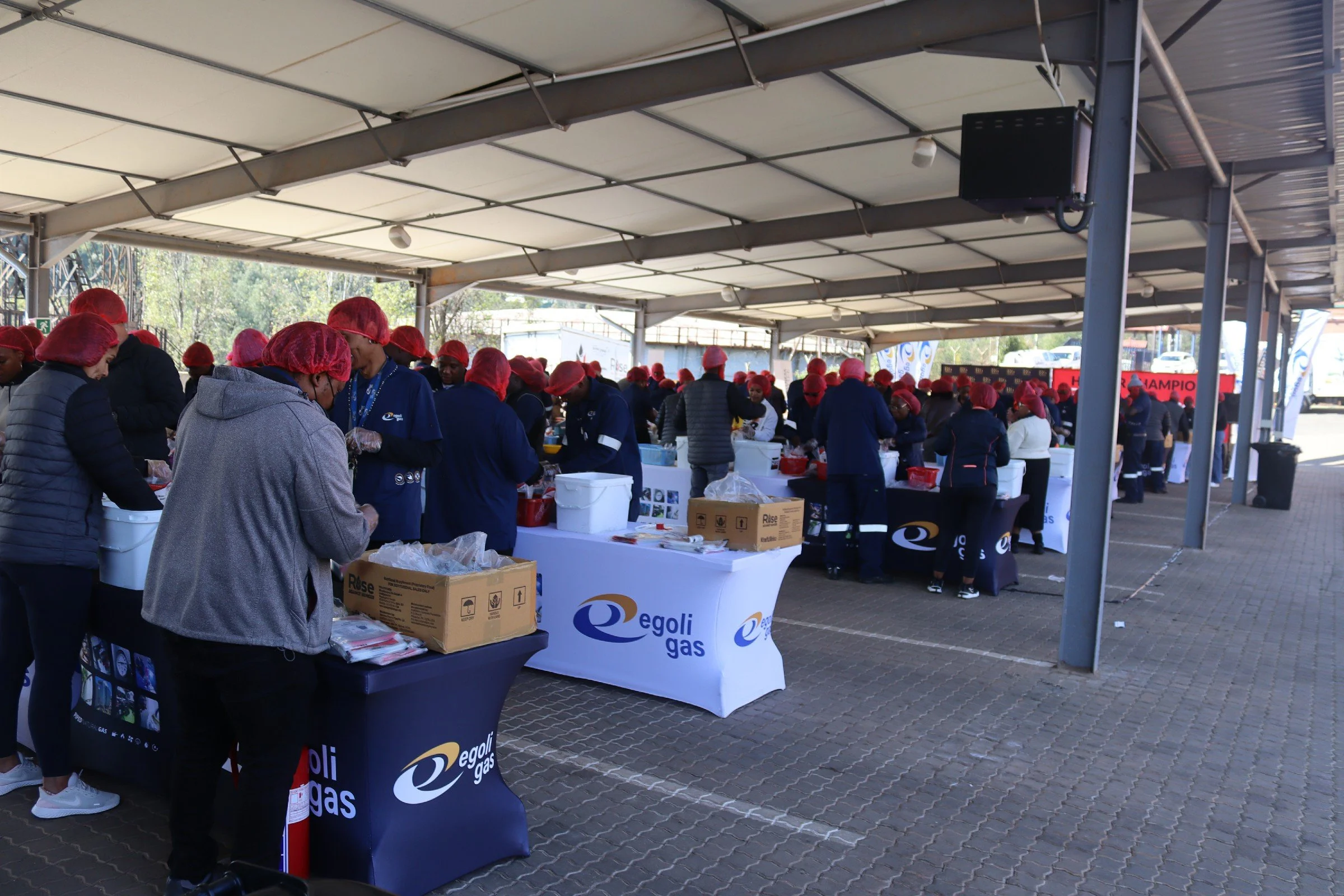 A large outdoor covered market or event space filled with many people wearing red hairnets, gathered around tables with supplies and food. The space has a metal roof with open sides, and some banners and signs are visible in the background.
