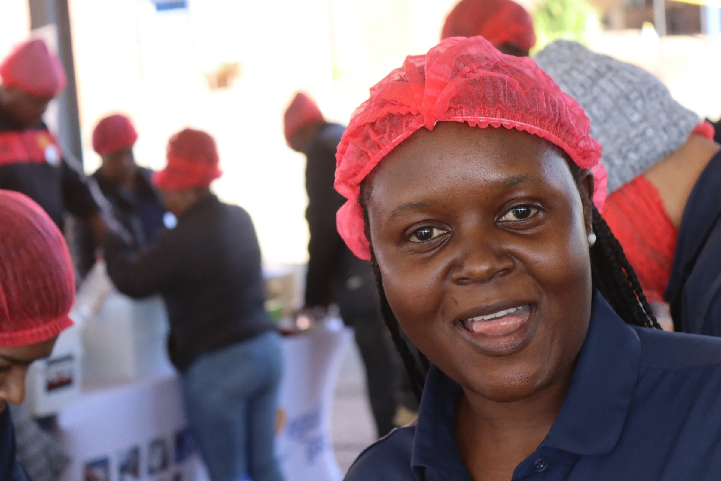 Close-up of a smiling woman with dark hair wearing a pink hairnet, surrounded by other women also wearing pink hairnets, indoors with natural light.