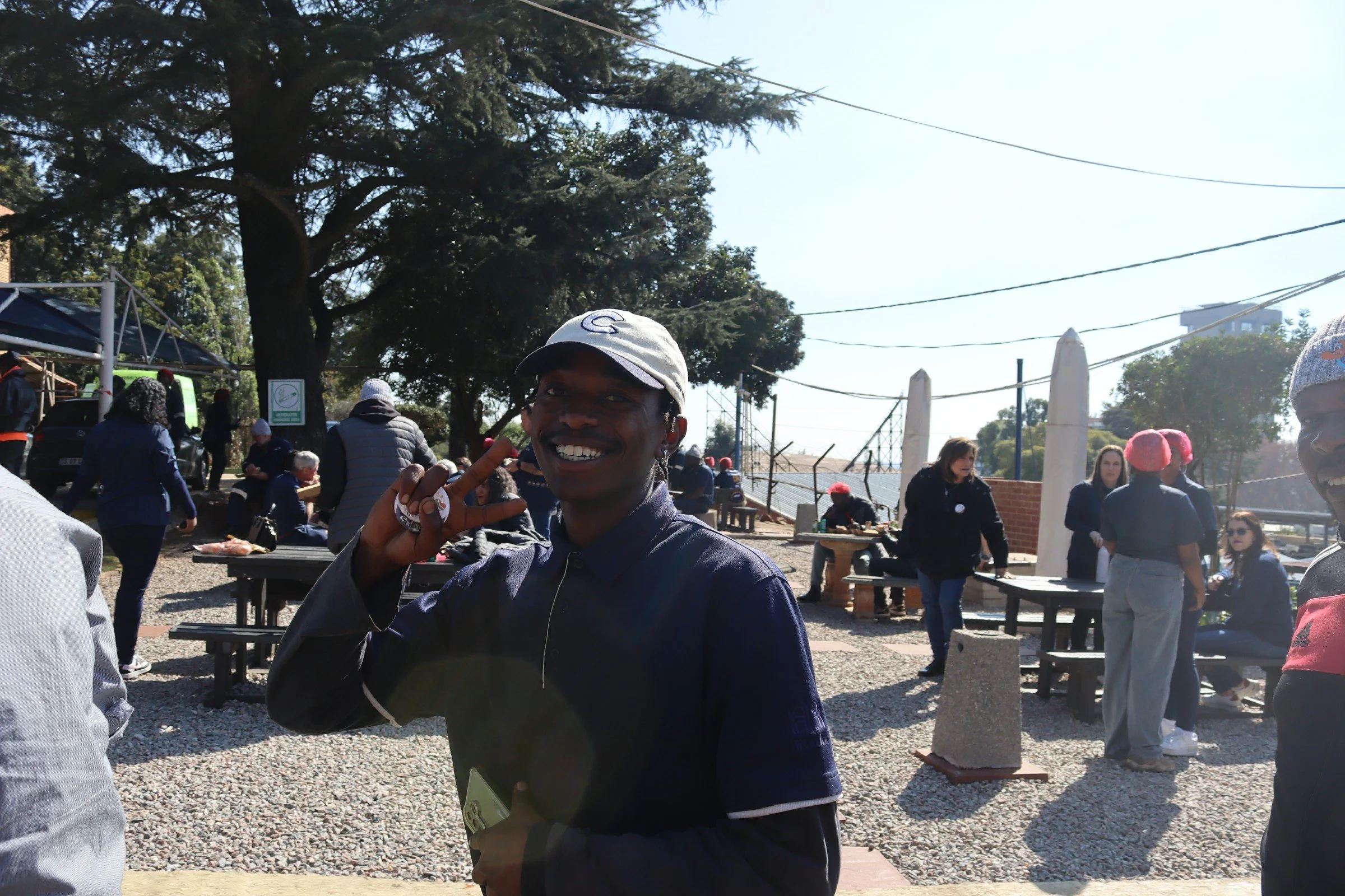 A group of people outdoors in a park or yard, with a young man smiling and making a peace sign near the camera, wearing a cap and dark jacket. In the background, some people are sitting at picnic tables and conversing, with others standing or walking