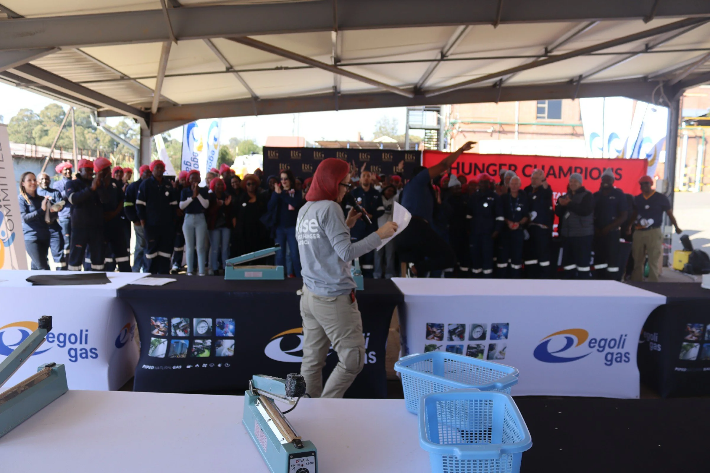 A group of people wearing red hats gather on a stage with a red banner reading 'HUNGER CHAMPIONS.' A woman in glasses speaks into a microphone, holding a paper. There are branded banners and tables with equipment in the foreground.