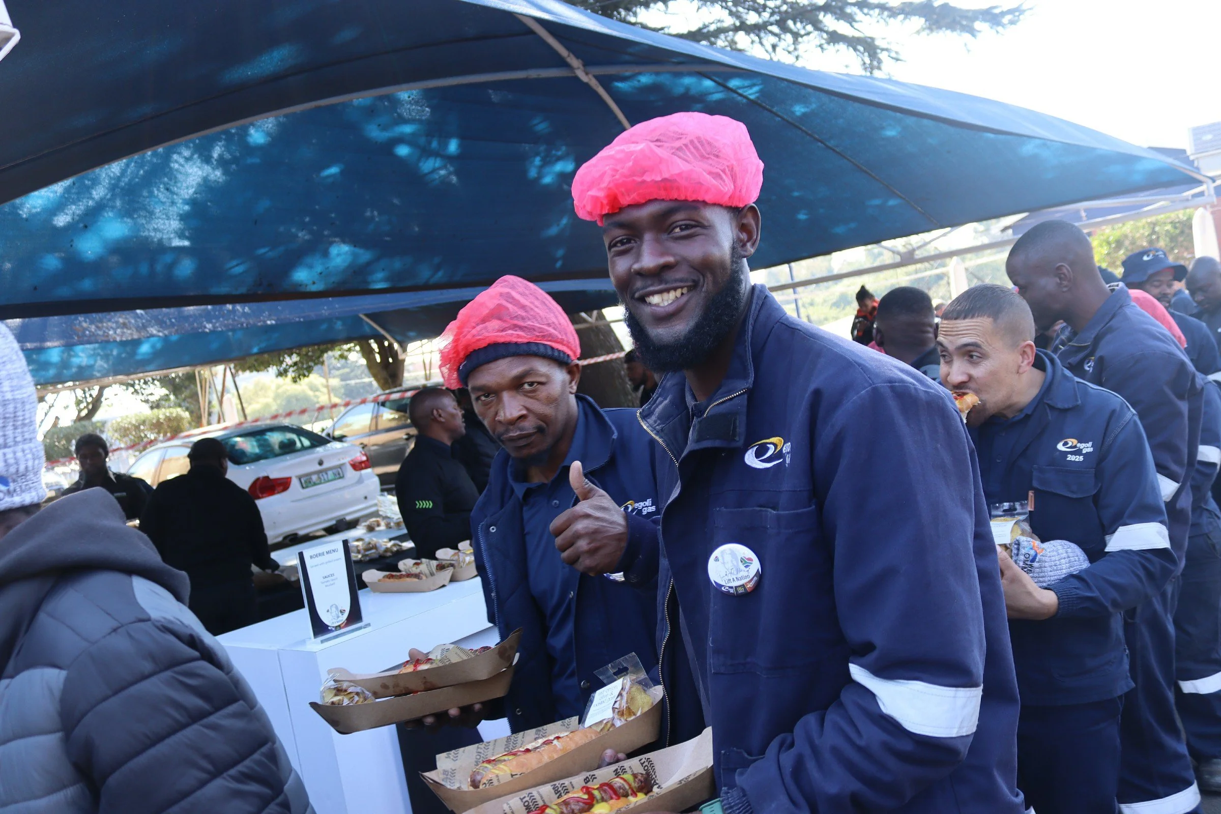 A group of people at an outdoor event, with two men in the foreground wearing matching navy jackets and pink hair covers, smiling and holding food packages. There are other people in the background, some with food, and a table with food items on it. 