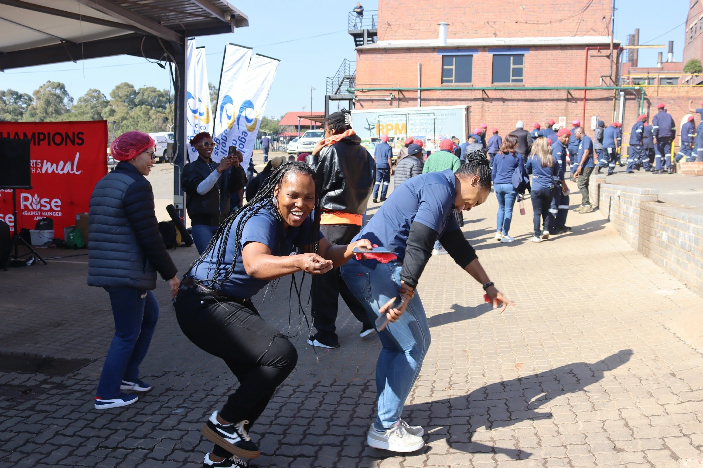 A group of people participating in an outdoor activity, holding onto a long ribbon, smiling, and laughing. In the background, there are more people in uniform walking and standing near a brick building and flags with logos, indicating a community eve