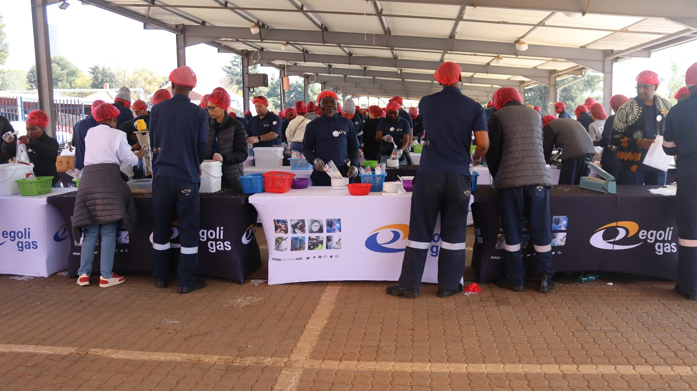 People wearing red hairnets working at a food service or distribution station under a large open-sided shelter, with tables and various containers, during daytime.