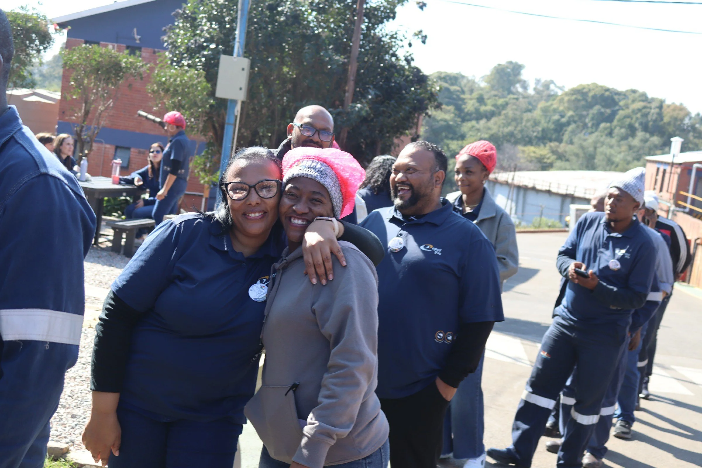 A group of smiling people standing outdoors in a line, with some hugging and others looking around, during daytime. They are wearing casual and matching navy shirts.