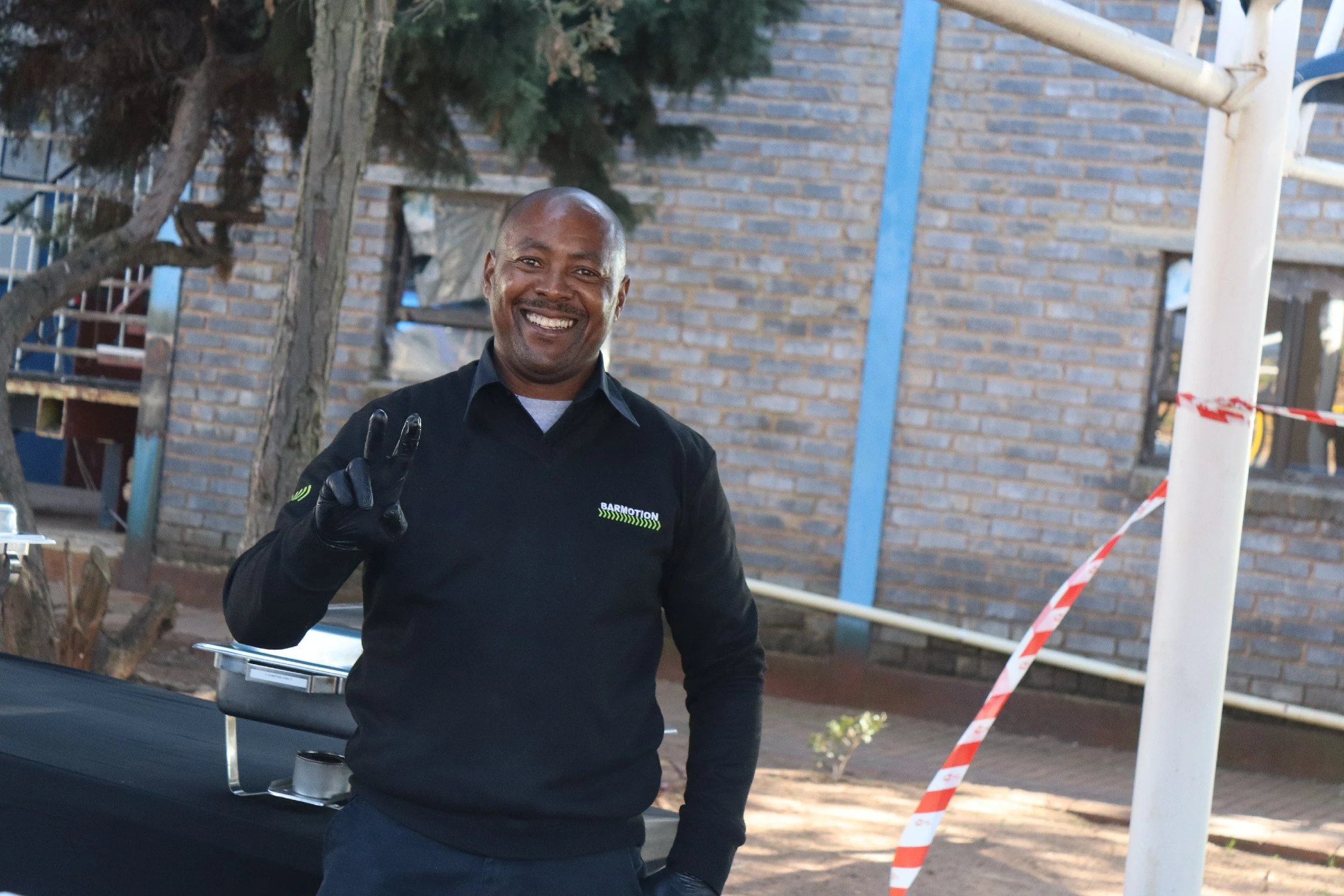 A smiling man with a shaved head, dressed in a black long-sleeve shirt, making a peace sign with his right hand, standing outdoors near a tree and a brick wall.