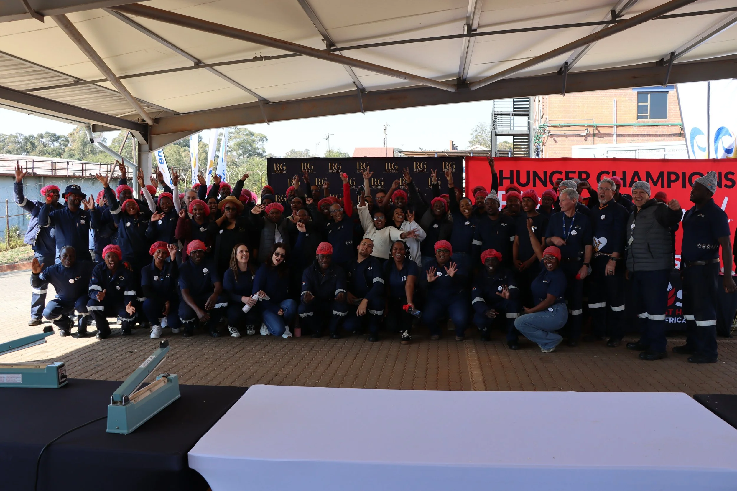 A large group of people wearing matching dark blue uniforms and red hairnets gathering outdoors on a stage with a banner that says 'HUNGER CHAMPIONS' and a backdrop with the words 'RG REBUILD'. They are smiling, raising their hands, and celebrating.