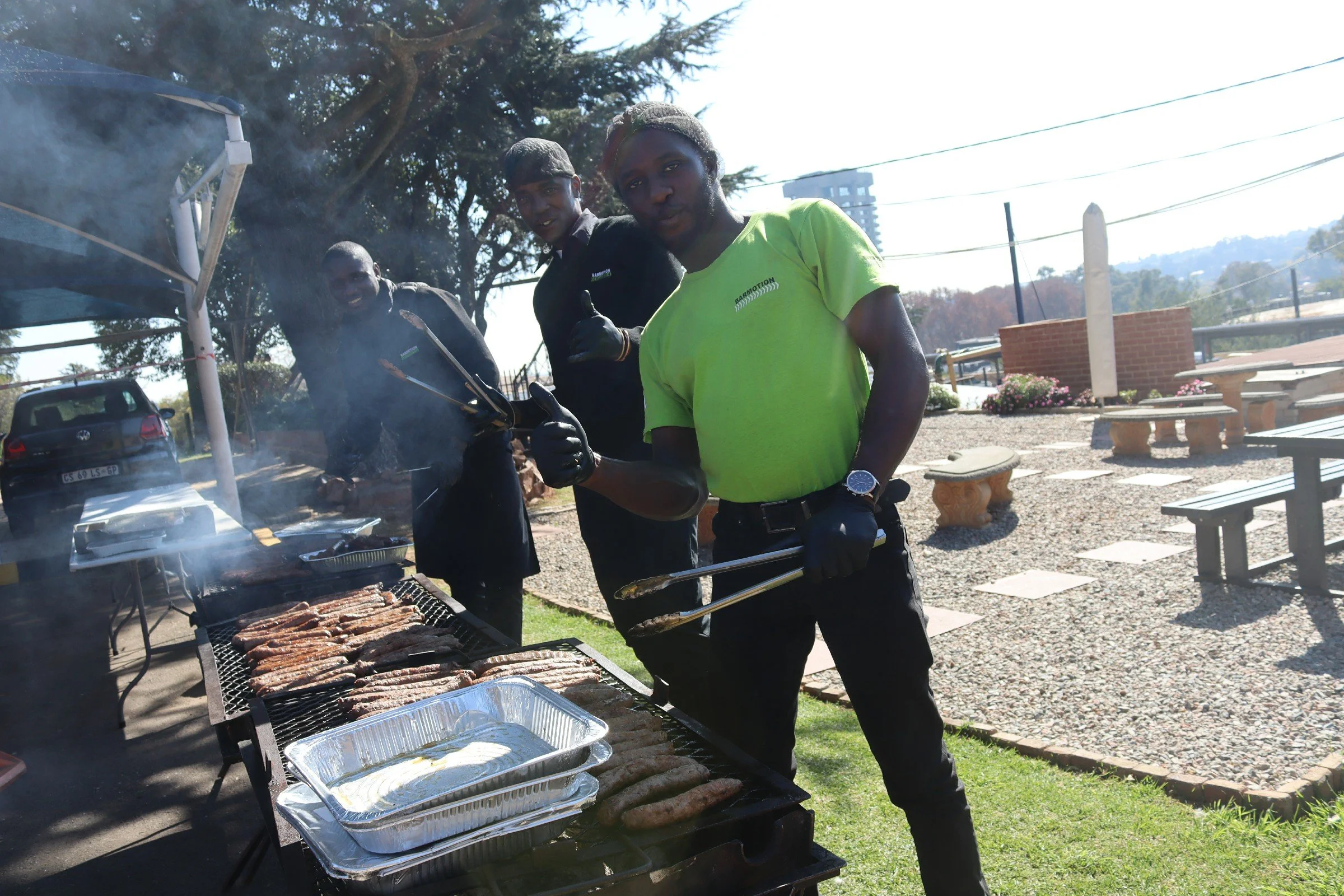 Four men are grilling meat outdoors, with two of them giving thumbs up. They are in a park-like setting with trees, benches, and a parking lot visible in the background. One man is wearing a bright green shirt, and the others are dressed in dark clot